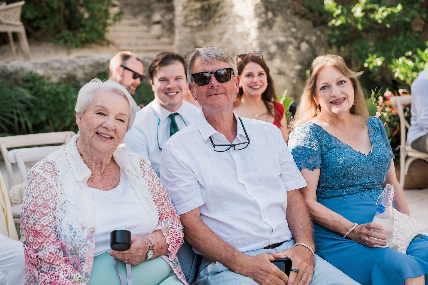 Smiling family group at an outdoor wedding celebration, featuring elegantly dressed guests posing together in a sunny garden.