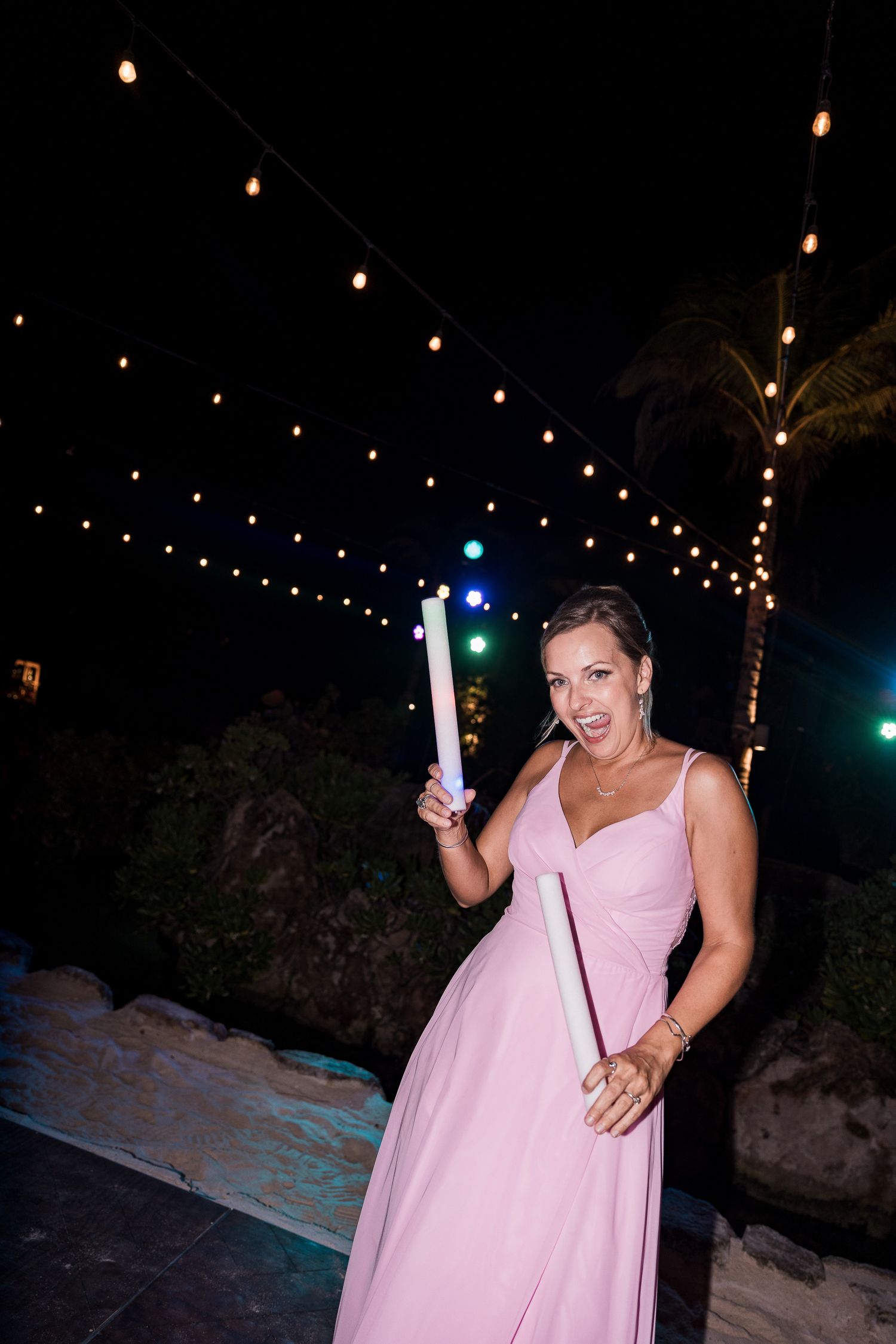 Woman in pink gown holding glow sticks at outdoor evening party with string lights.