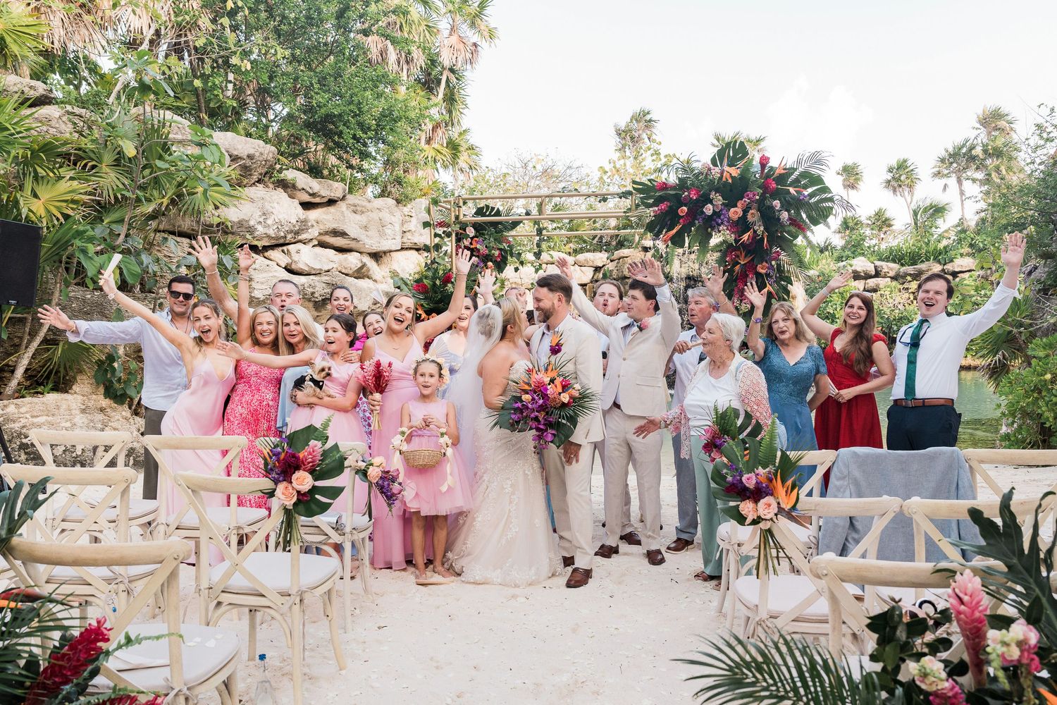 Joyful wedding party celebrating outdoors at a tropical venue with lush greenery and colorful floral arrangements.