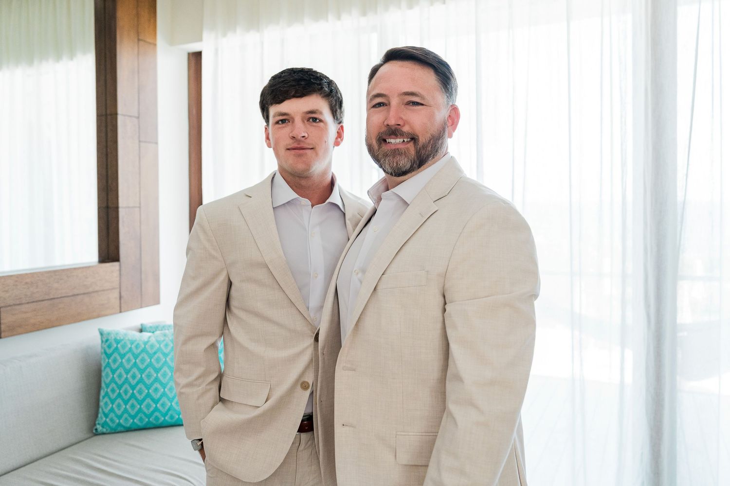 Two men in matching beige suits smiling together indoors near white curtains and a teal decorative pillow.