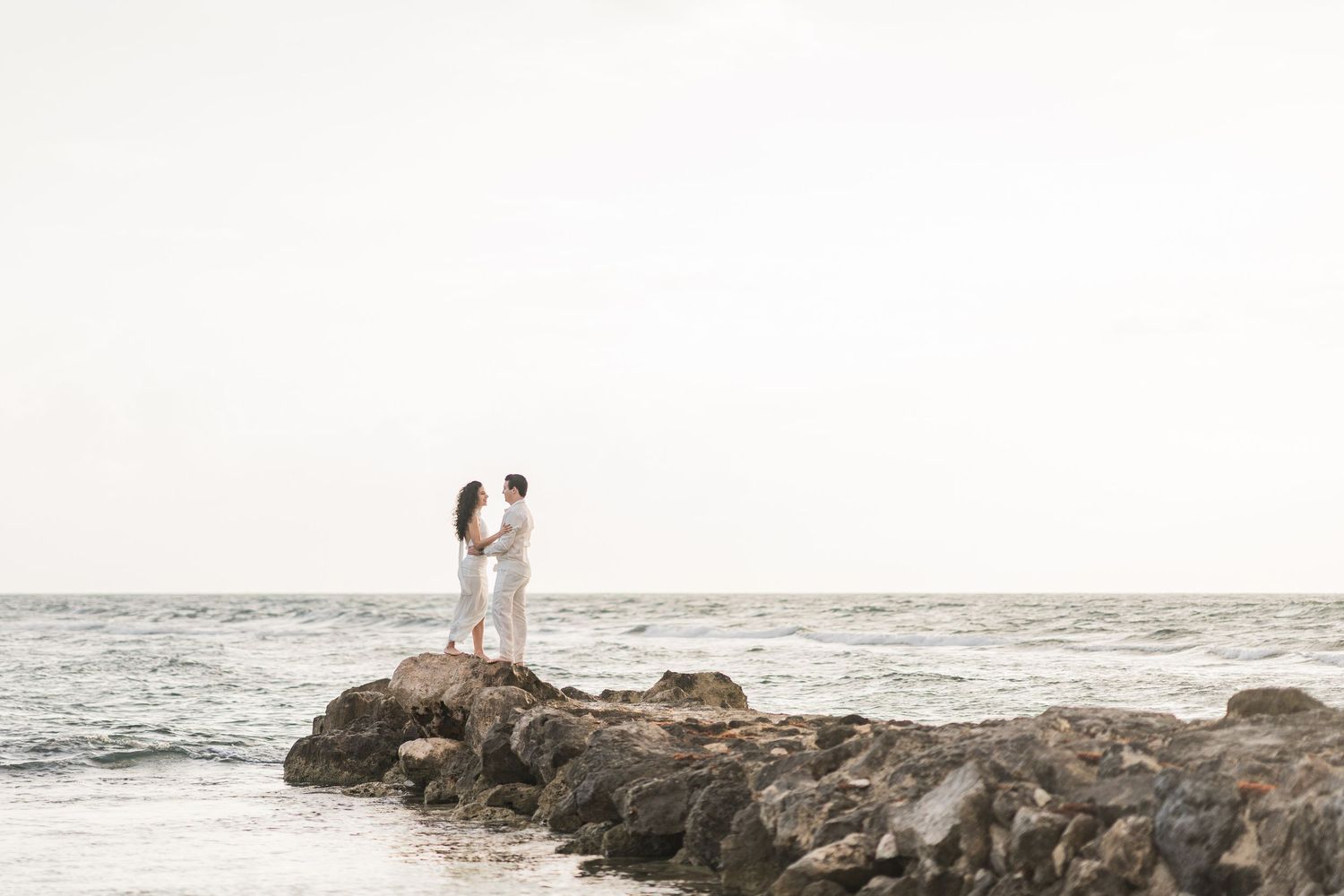 Couple in white outfits standing on rocky jetty by ocean shore at sunset.