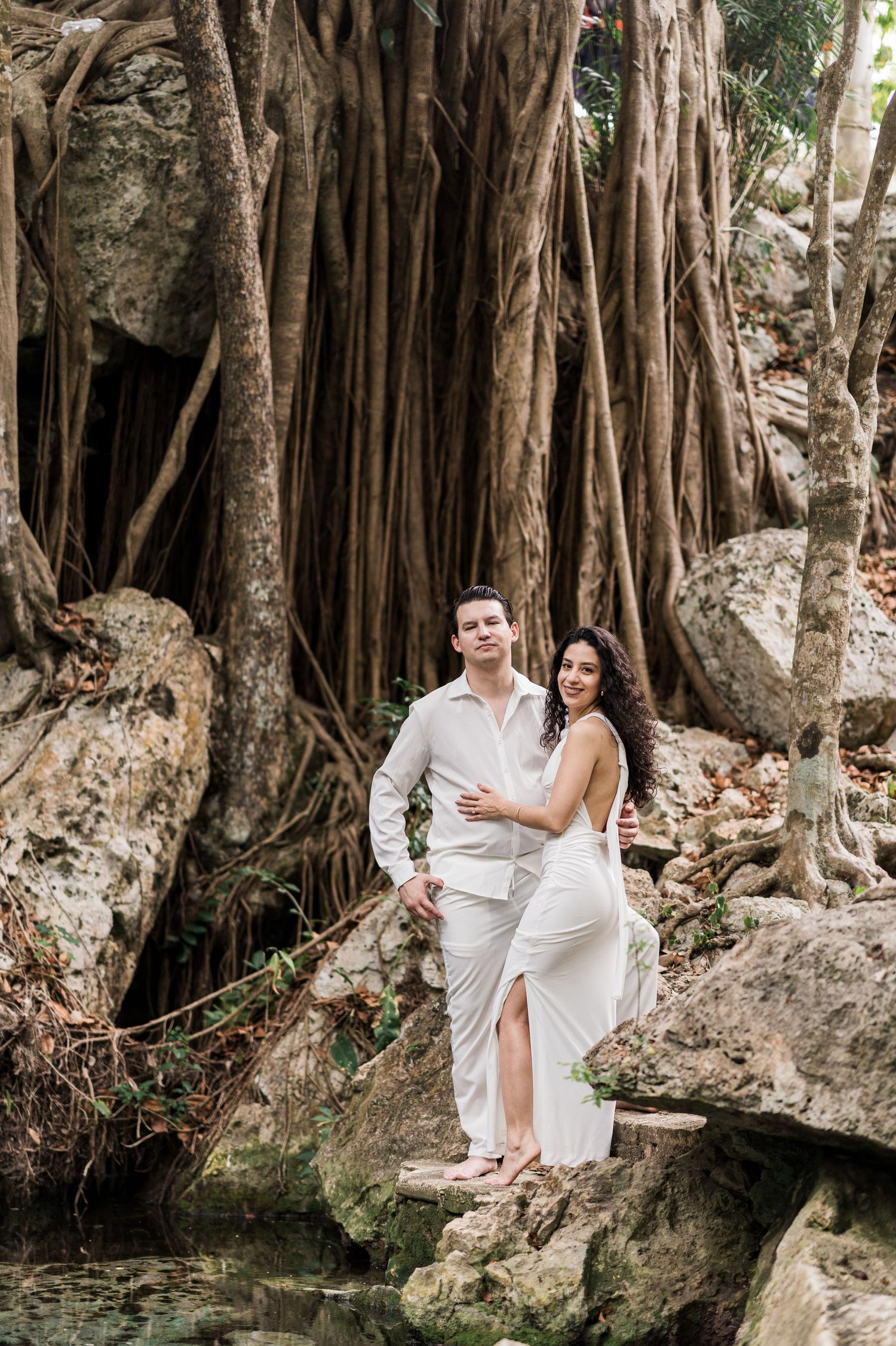 Couple in white outfits posing on rocks before large banyan tree roots in a tropical setting.