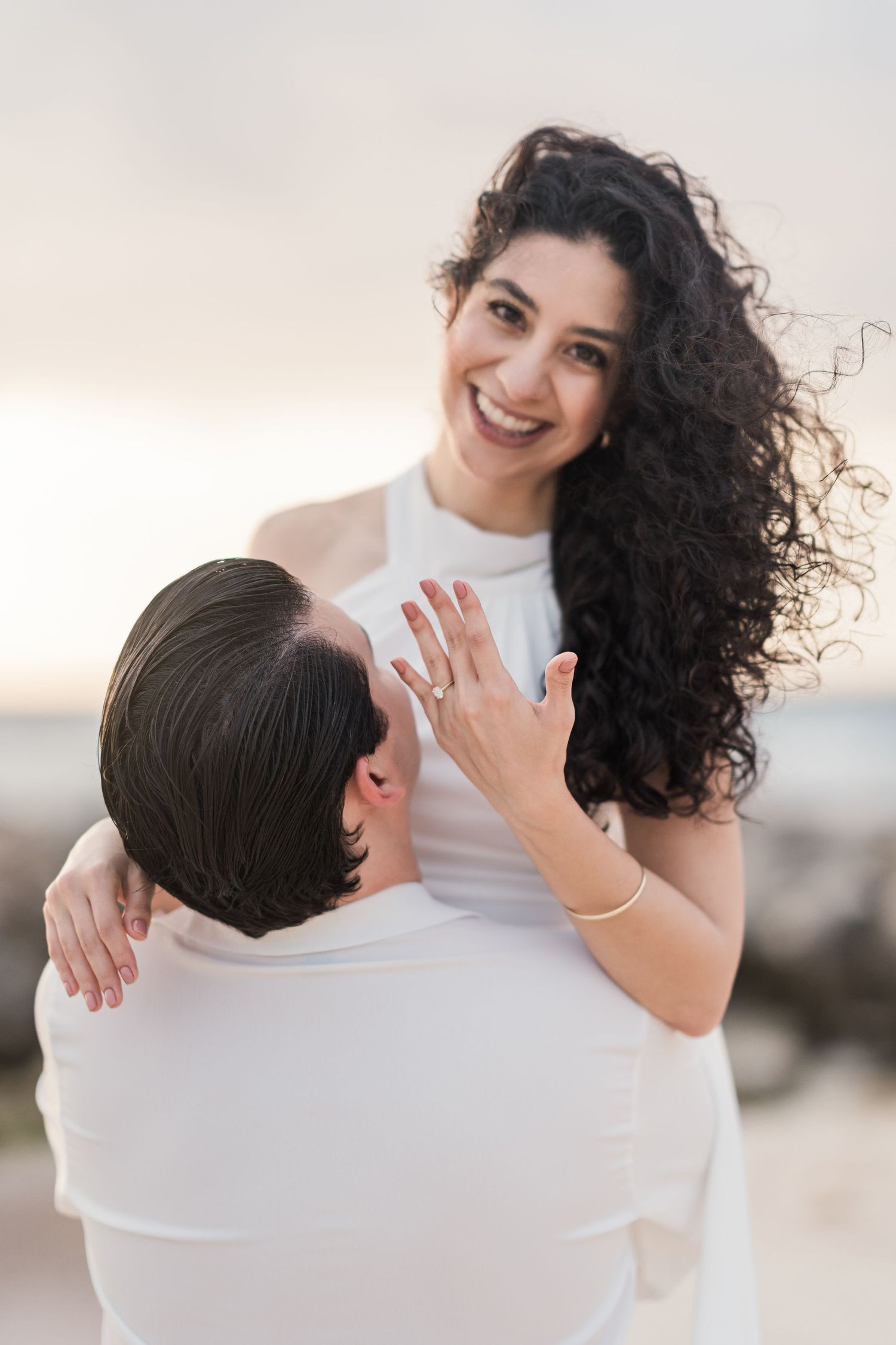 A joyful woman in white shows her engagement ring while being lifted by her partner on a beach at sunset.