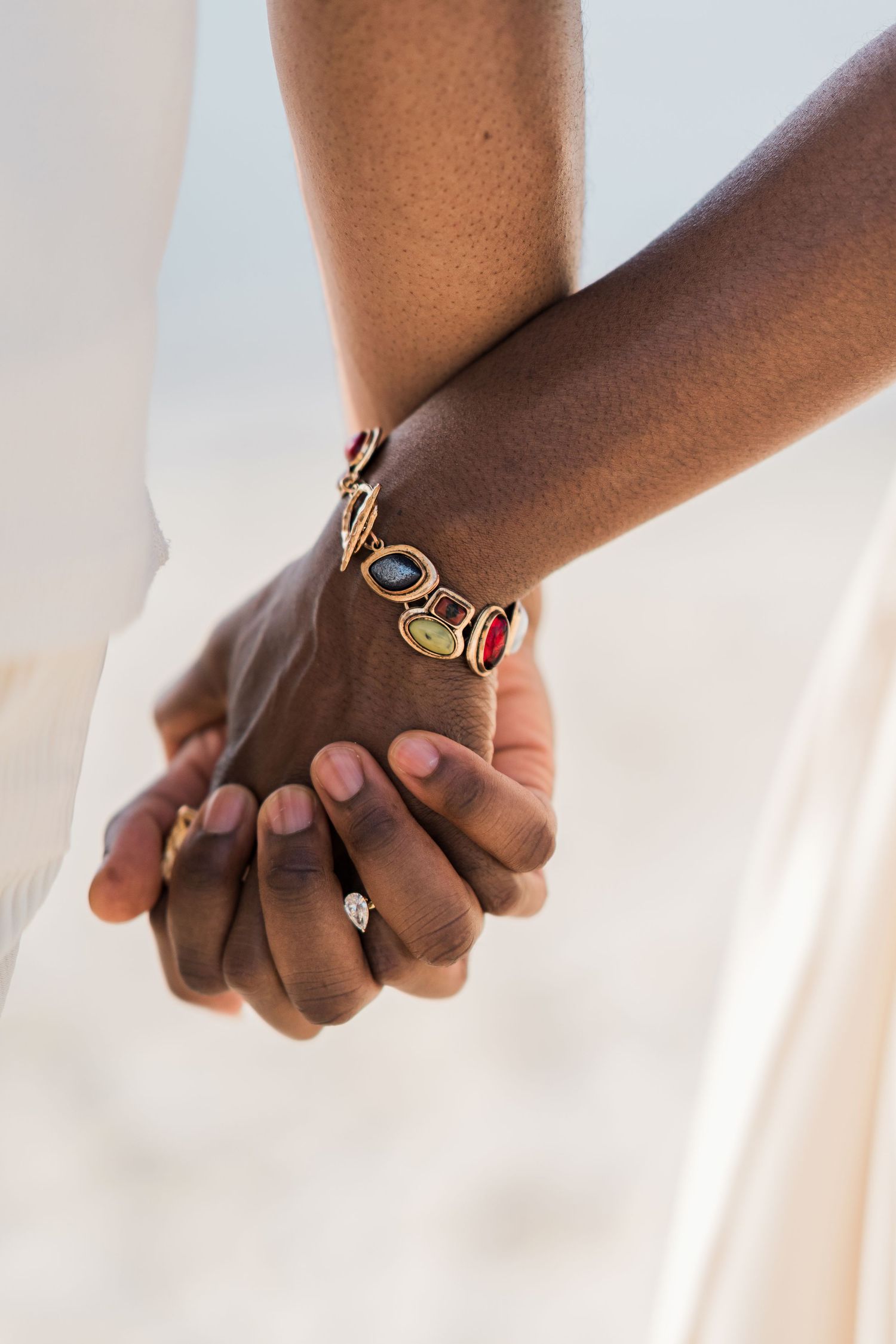 Close-up of two people holding hands on a beach, woman wearing a colorful gemstone bracelet and diamond engagement ring.