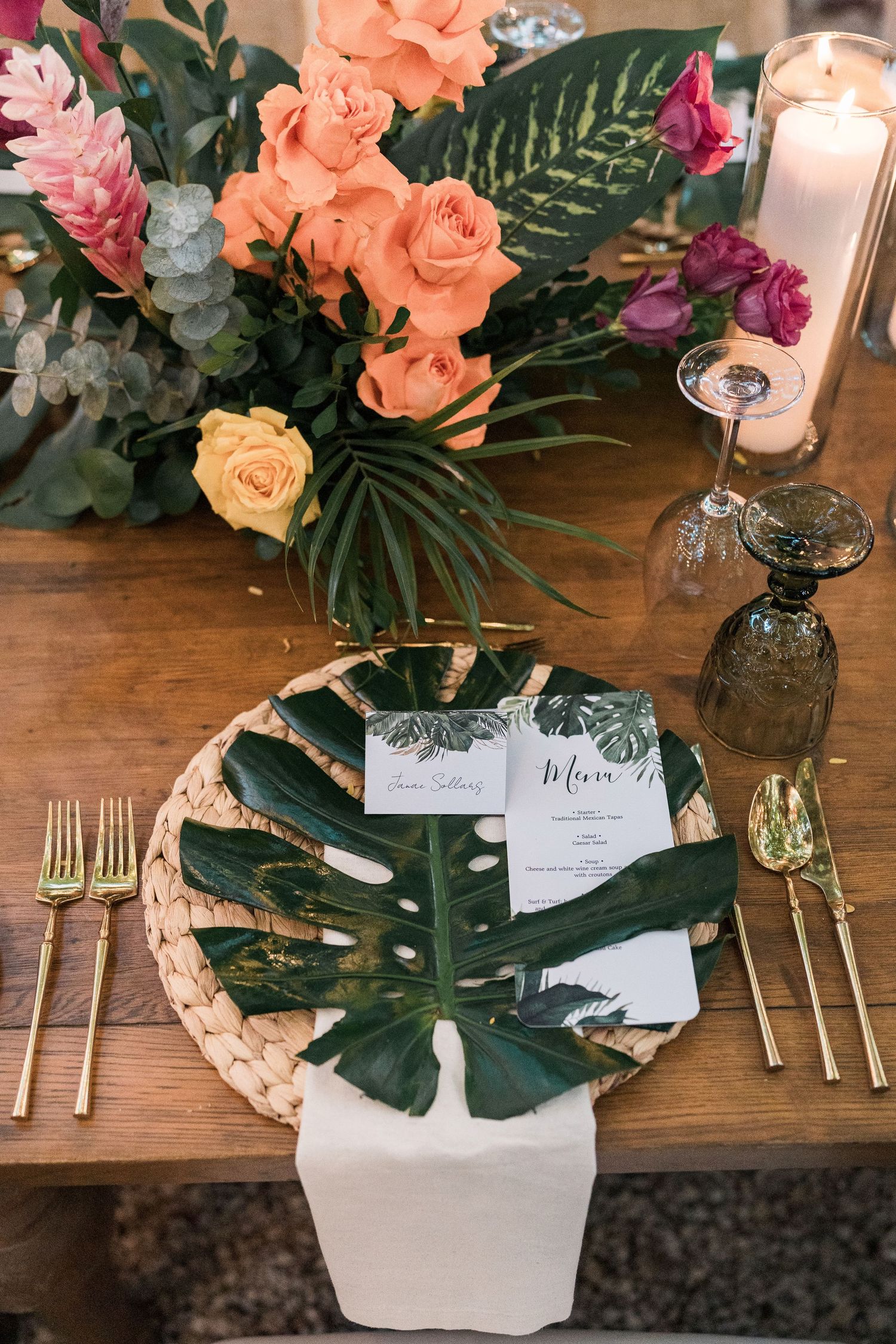 Secrets Akumal Wedding - Tropical wedding tablescape with monstera leaf plate, gold cutlery, woven charger, and vibrant floral centerpiece.