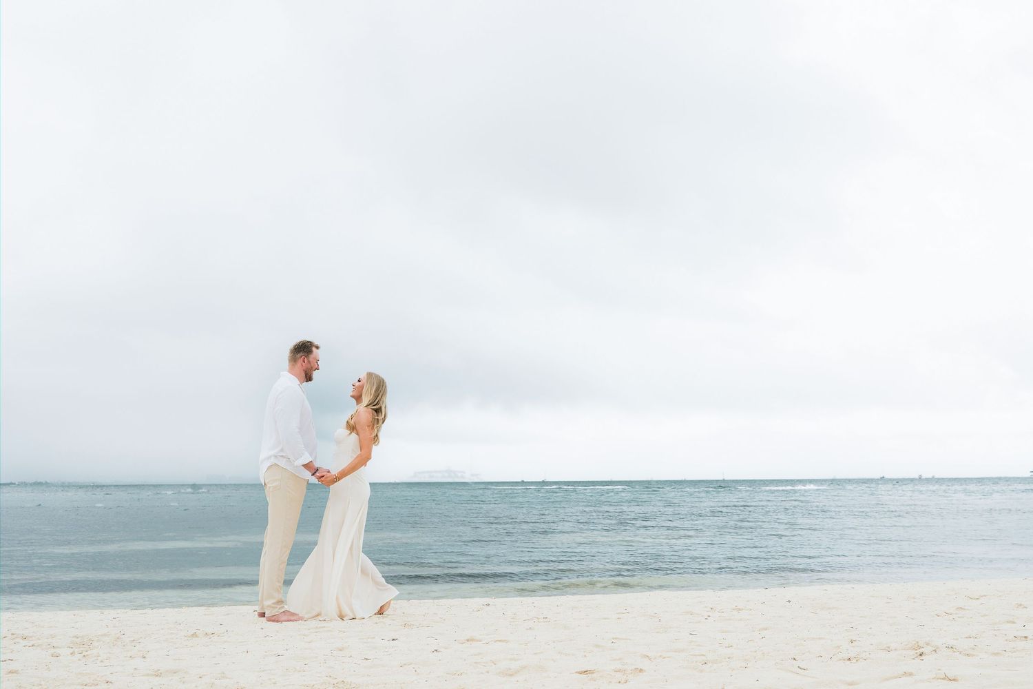 CANCÚN ENGAGEMENT PHOTOSHOOT - Couple holding hands on a white sand beach with calm ocean waters and overcast sky in a romantic setting.