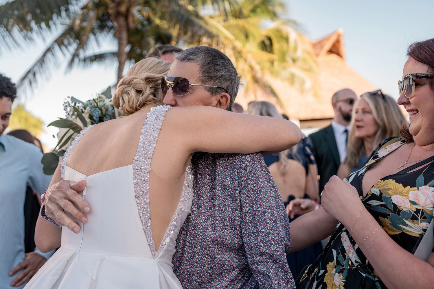 Bride in white gown hugging a guest in sunglasses at an outdoor beach wedding ceremony.