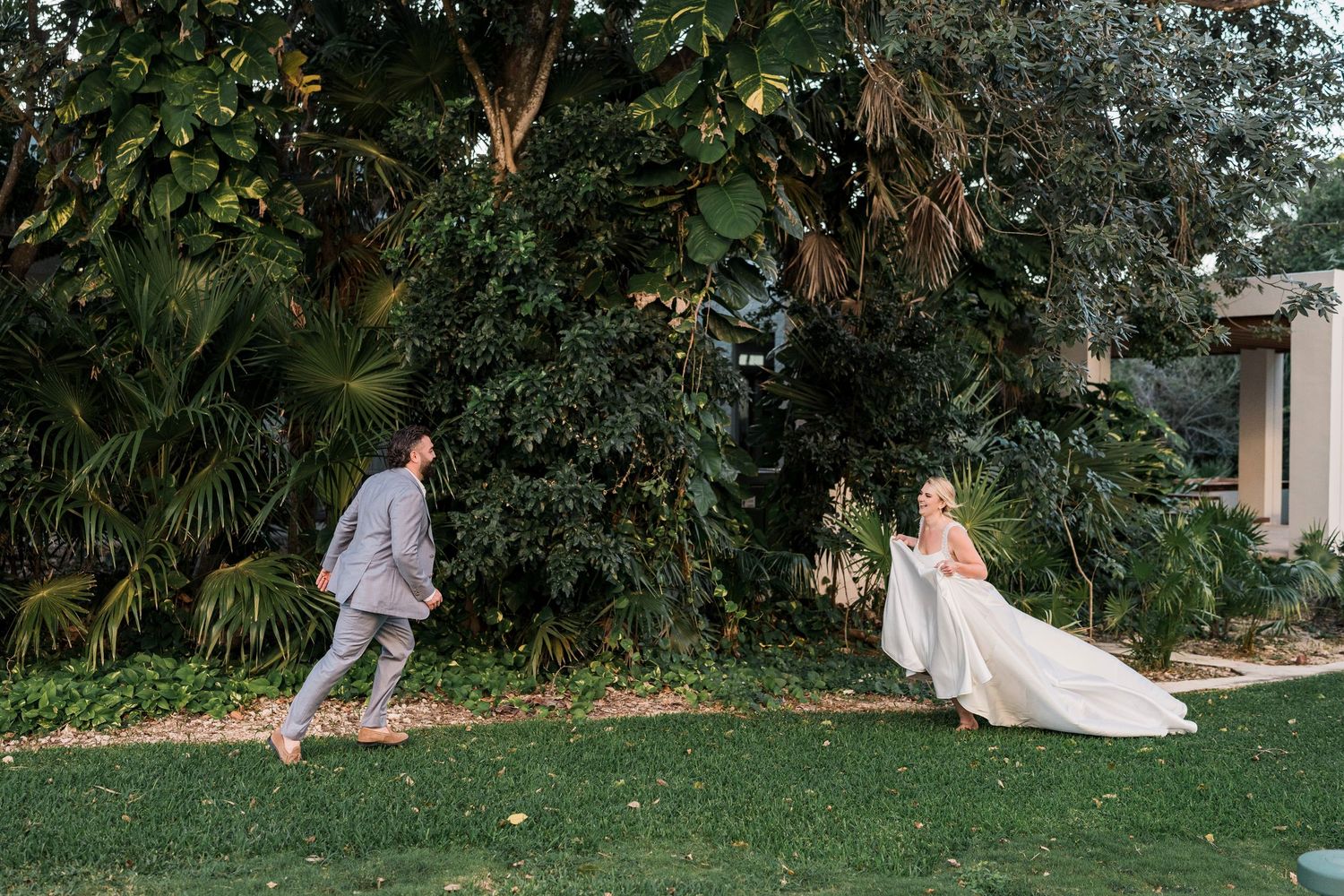 Bride and groom sharing a joyful outdoor moment on lush green lawn surrounded by tropical trees.