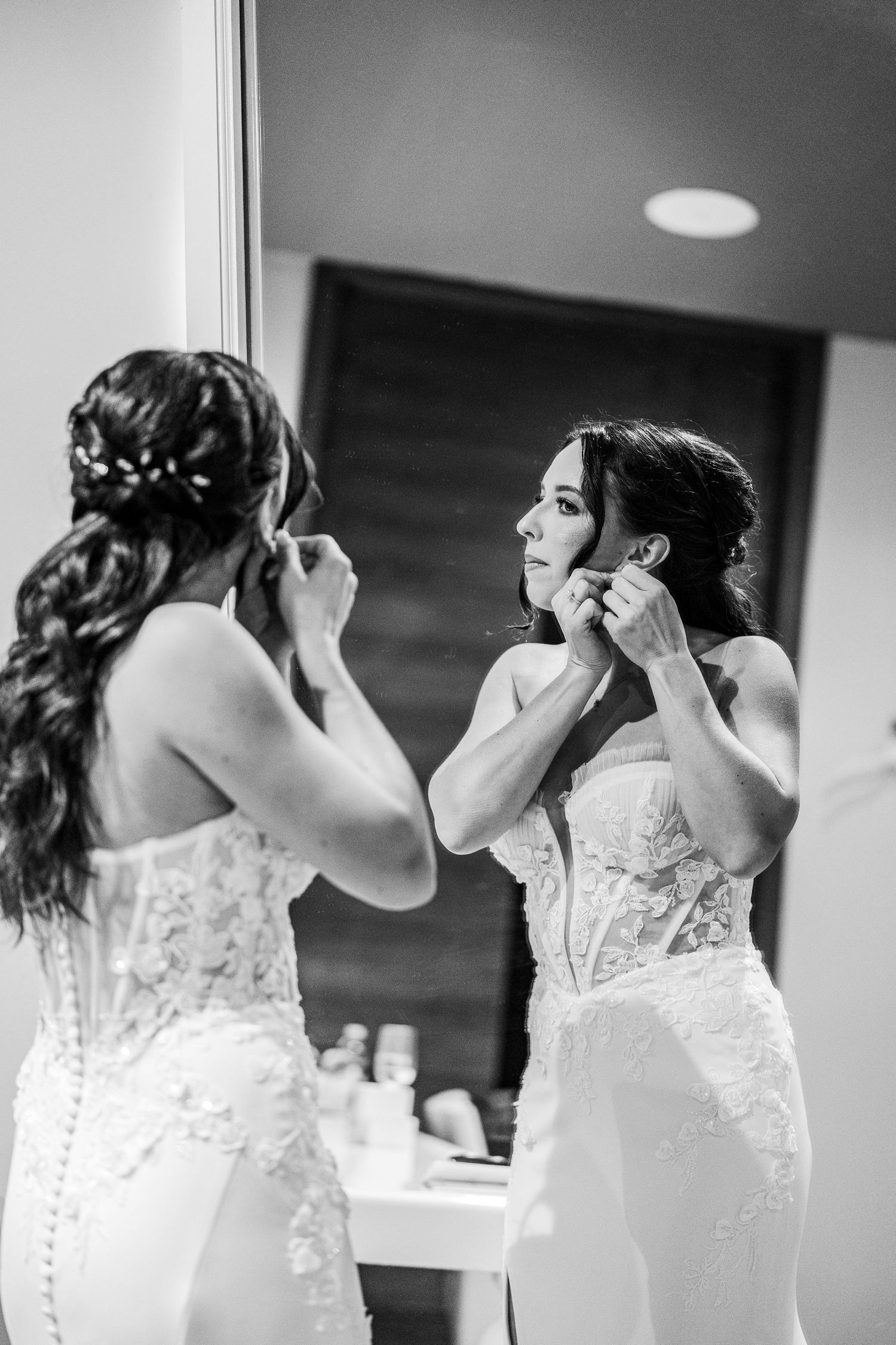 Bride in lace wedding gown putting on earrings while looking in mirror, black and white wedding photo.