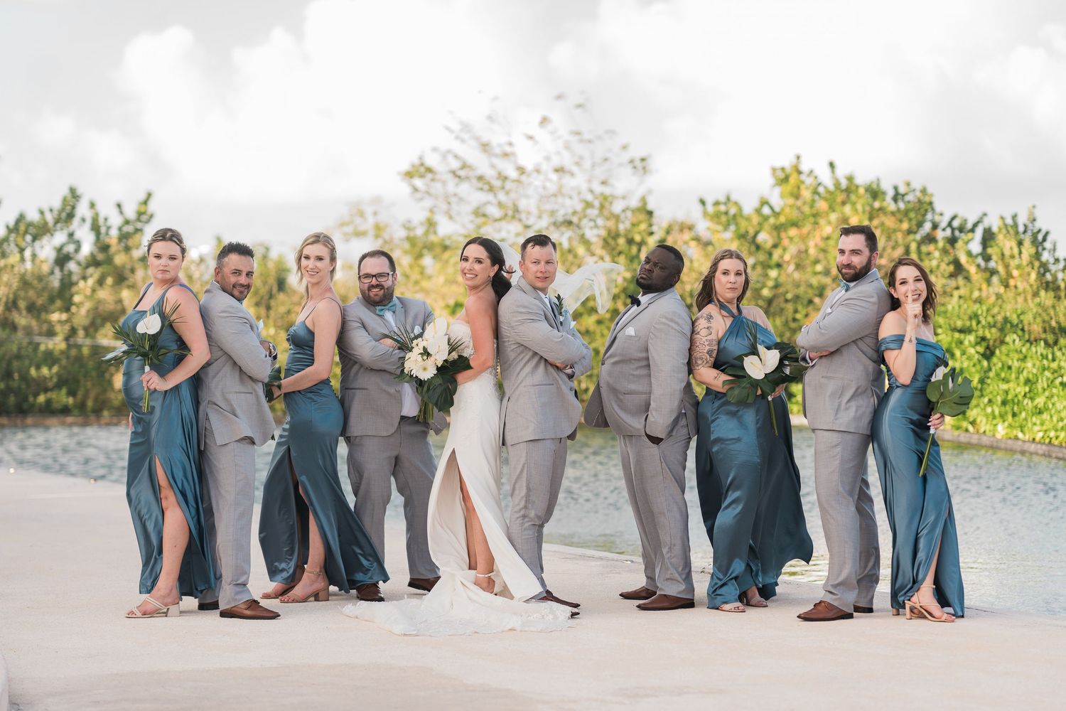 Wedding party posing outdoors near a pool, bridesmaids in blue dresses and groomsmen in gray suits flanking the bride and groom.
