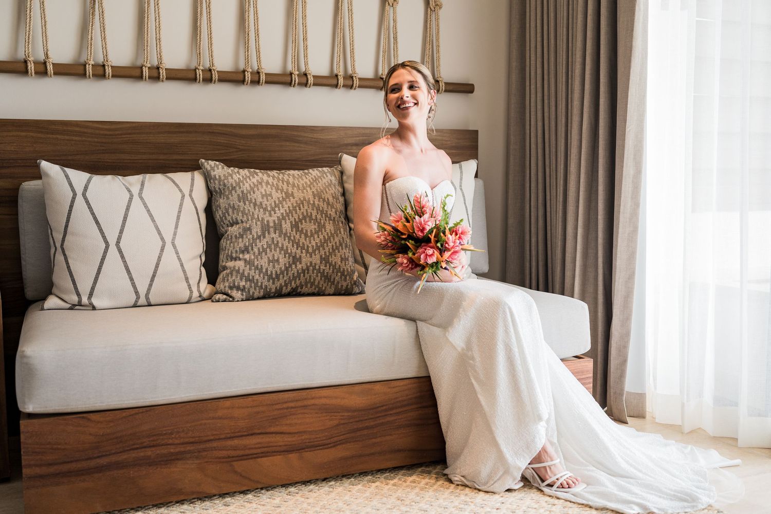 Bride in white gown holding tropical bouquet sits on wooden bed frame in elegant hotel room.