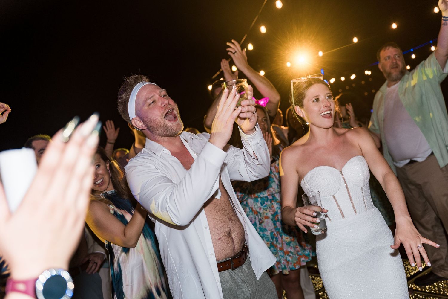Bride in strapless white gown and groom in white celebrating joyfully at outdoor wedding reception at night.