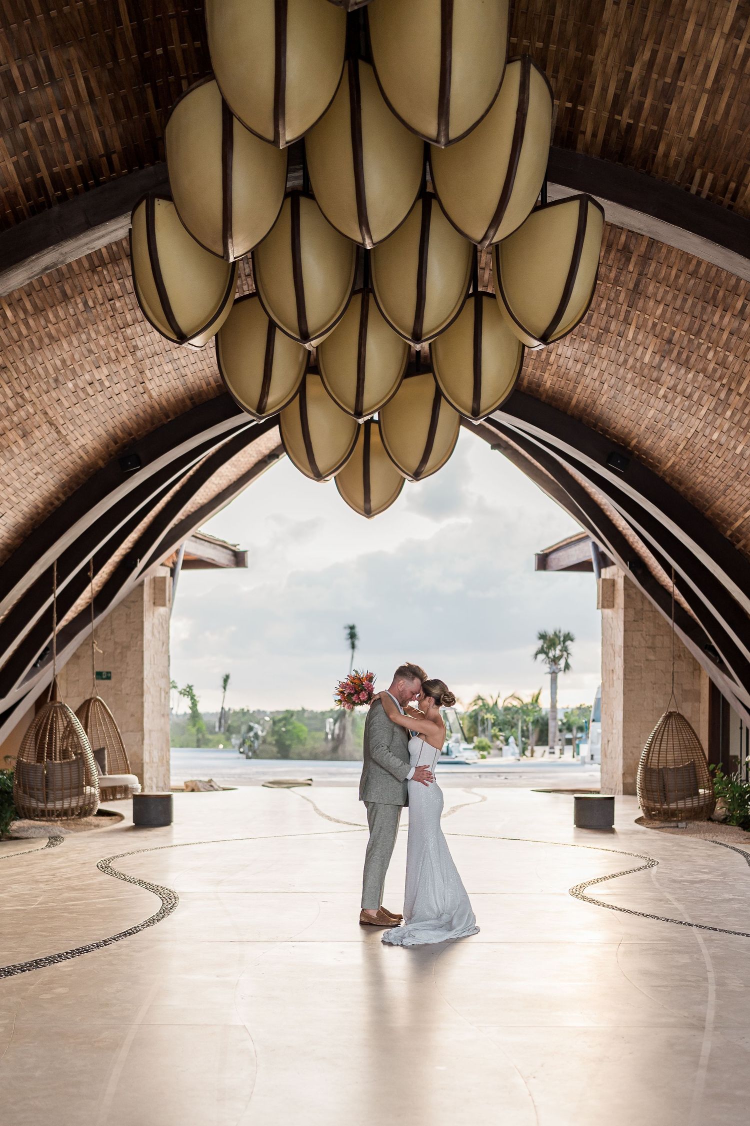 Bride and groom embrace under stunning architectural arch with decorative ceiling at tropical wedding venue.