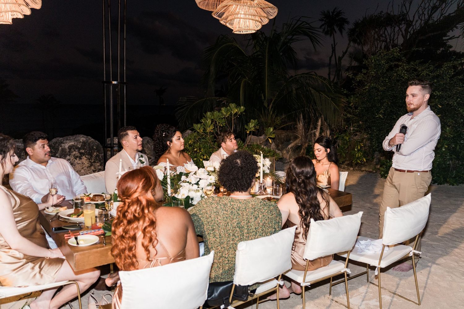 Xcaret Arte Wedding - A man gives a toast at an elegant outdoor dinner party at night with guests seated at a decorated table.