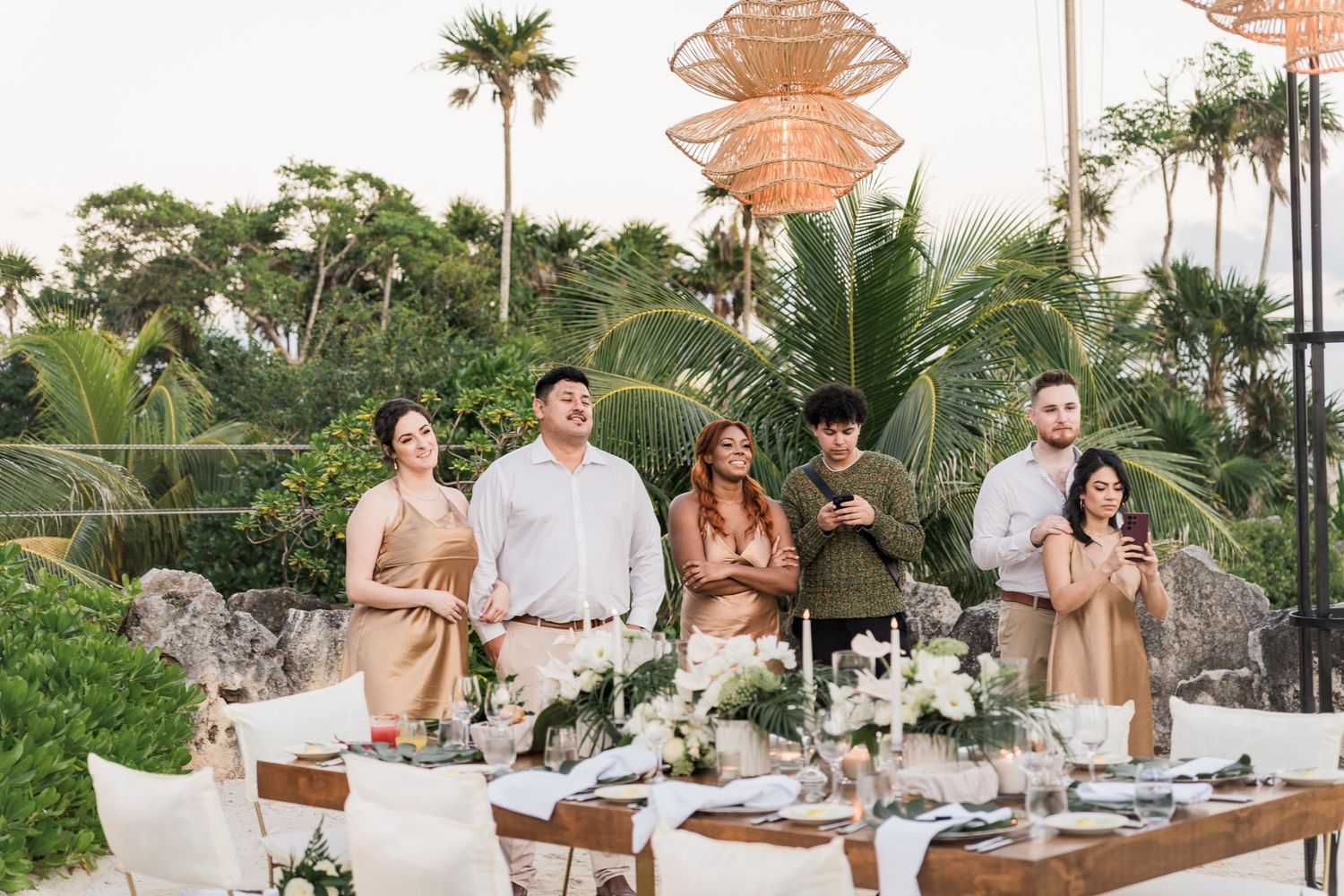 Xcaret Arte Wedding - Guests gather around an elegant outdoor dining table with floral arrangements, palm trees, and rattan pendant lights