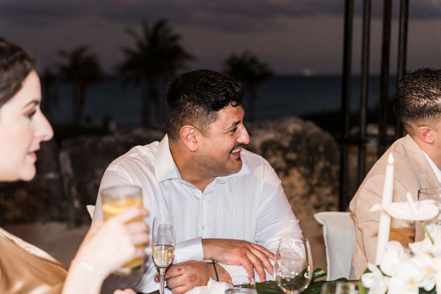 Xcaret Arte Wedding - Man in white shirt smiling at outdoor evening wedding reception table with champagne glasses and ocean view.
