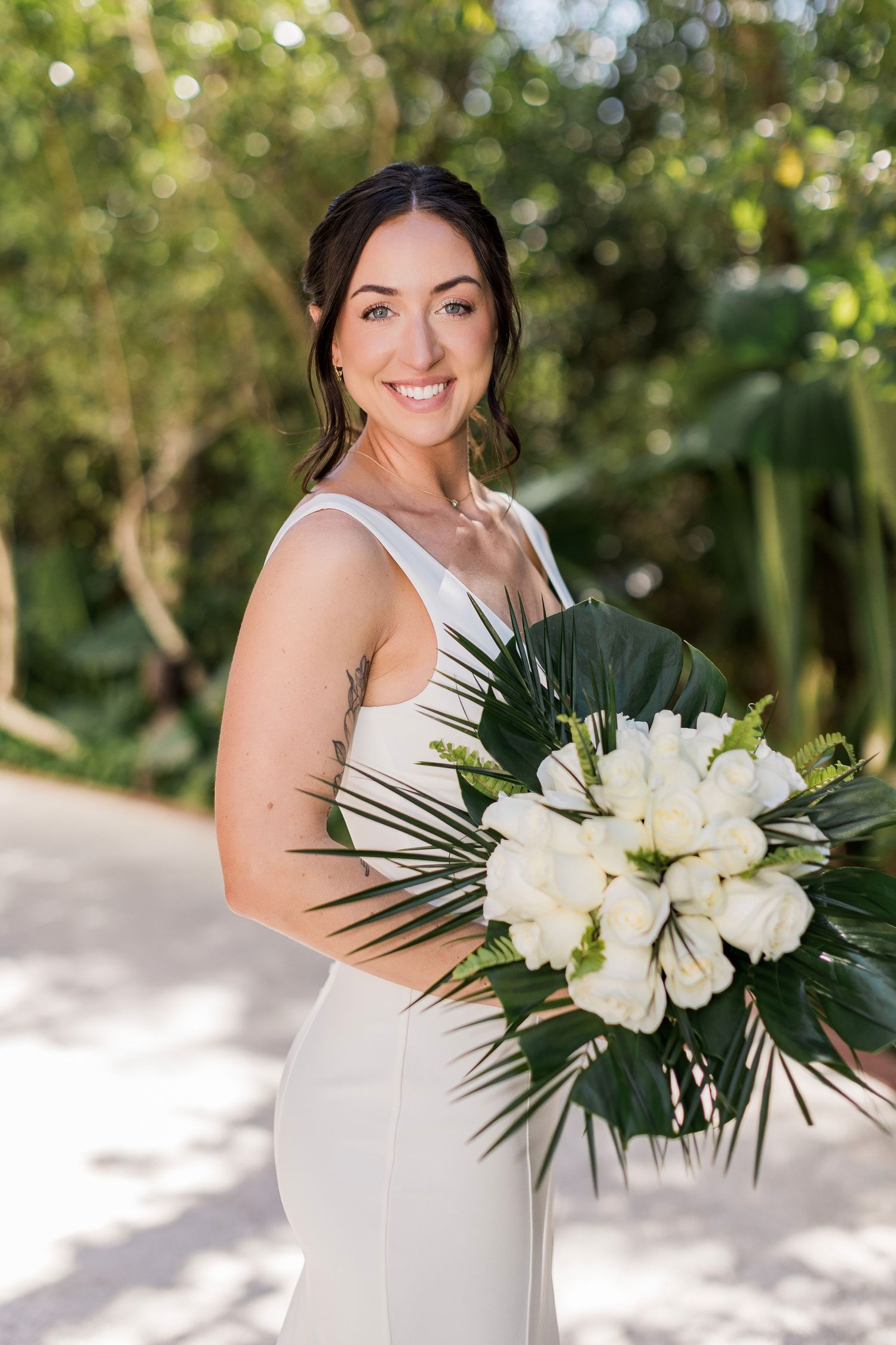 Smiling bride in white gown holding tropical bouquet of white roses and palm leaves outdoors.