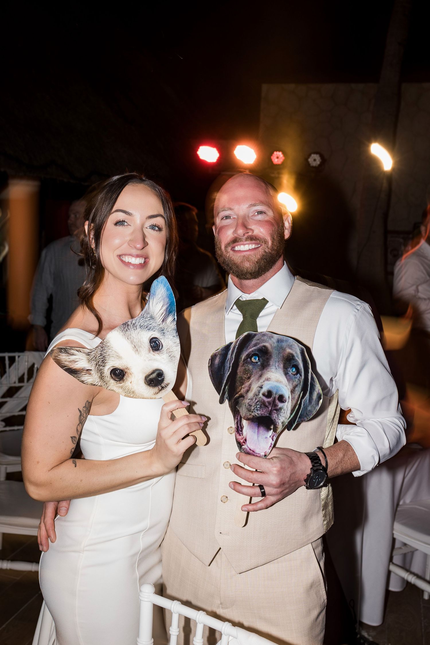 Newlywed couple smiling at wedding reception holding oversized dog face photo props at night.