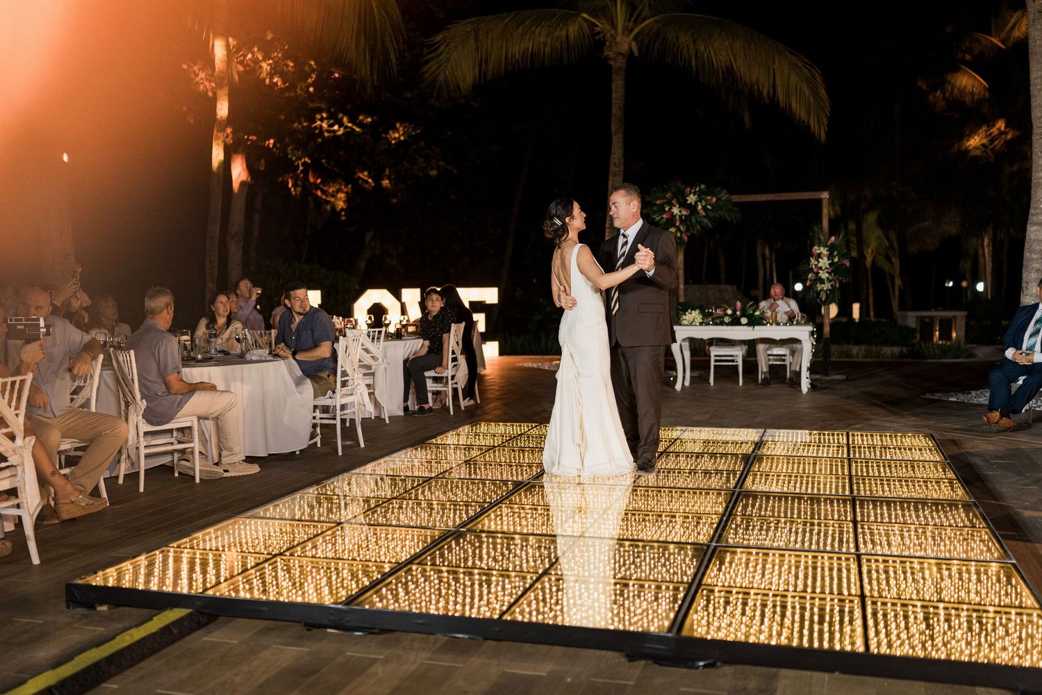 Bride and groom share first dance on illuminated gold dance floor at outdoor evening wedding reception.