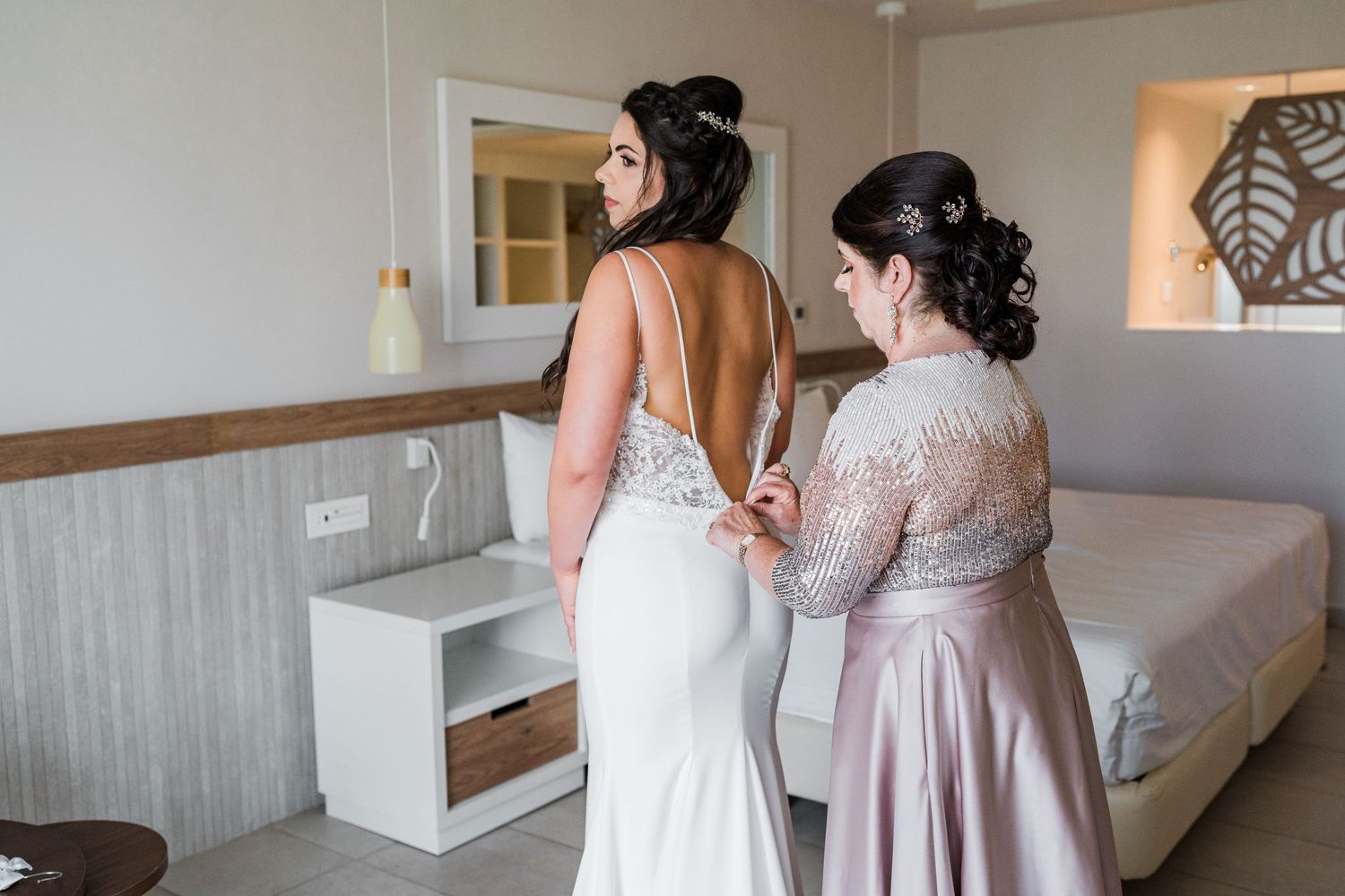 Bridesmaid fastening back of brides fitted white wedding gown in bright hotel room.
