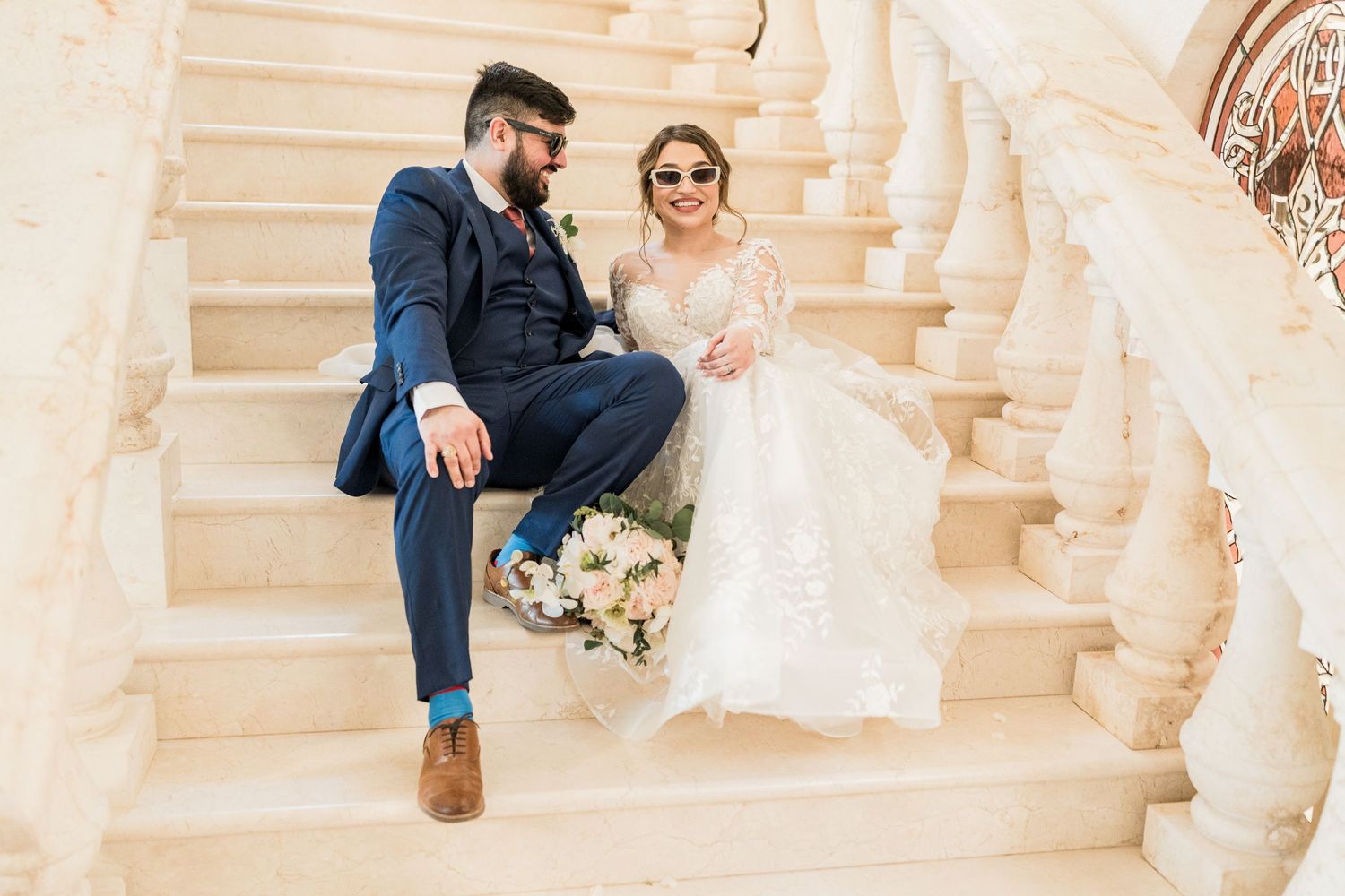 Bride and groom sit together laughing on elegant marble staircase with floral bouquet.