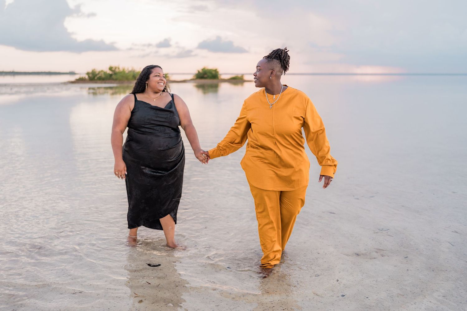 Two women holding hands walking through shallow ocean water at sunset.