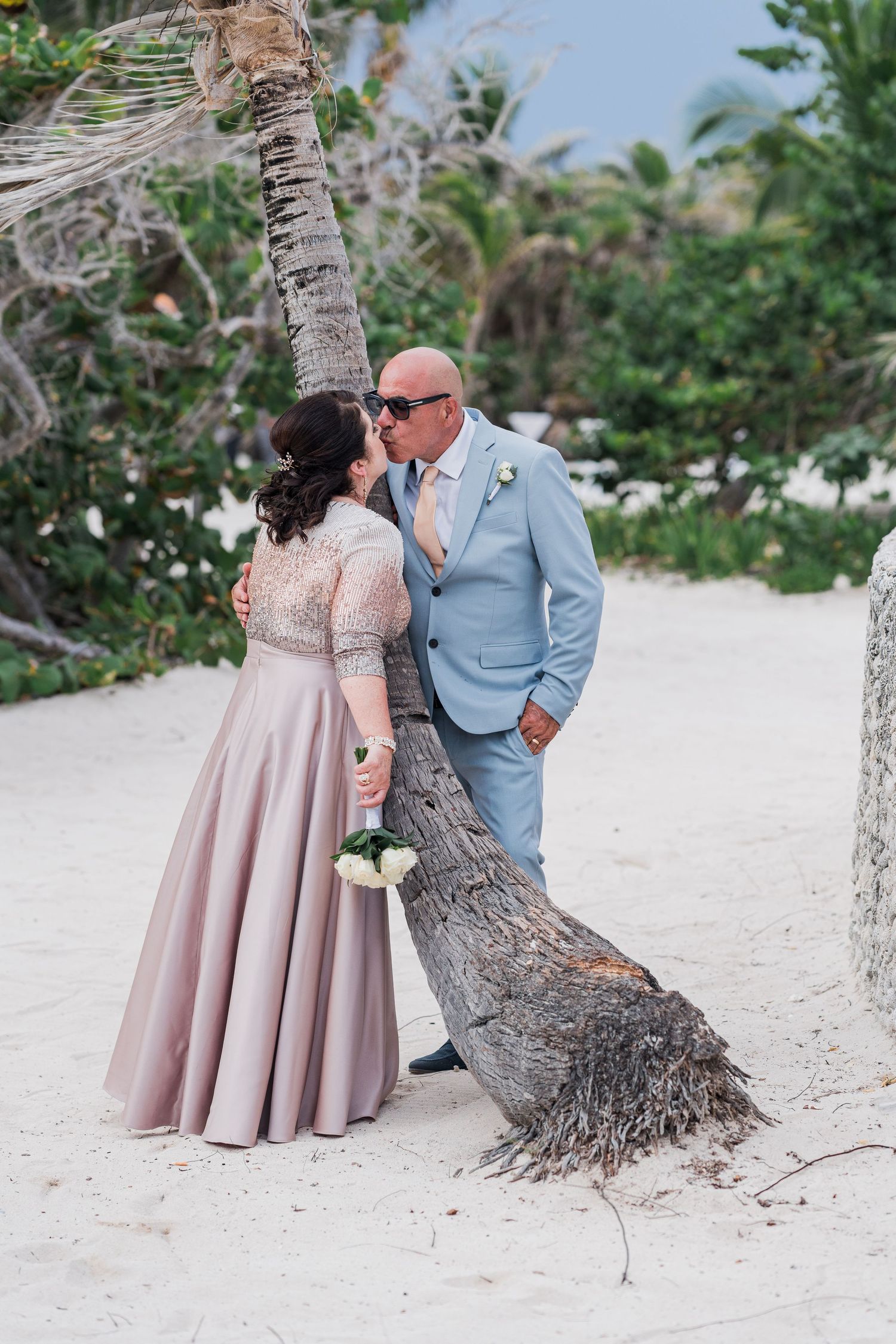 Couple sharing a romantic kiss beside a leaning palm tree on a white sandy beach.