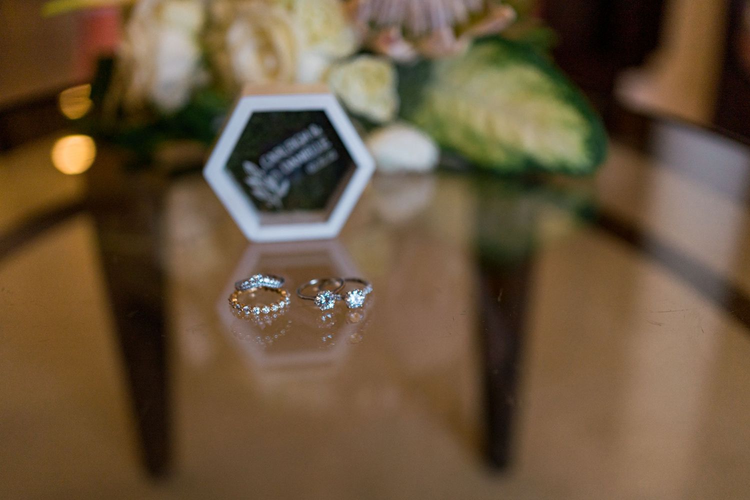 Close-up of elegant diamond wedding rings on a glass surface with a floral bouquet and ring box in the background.