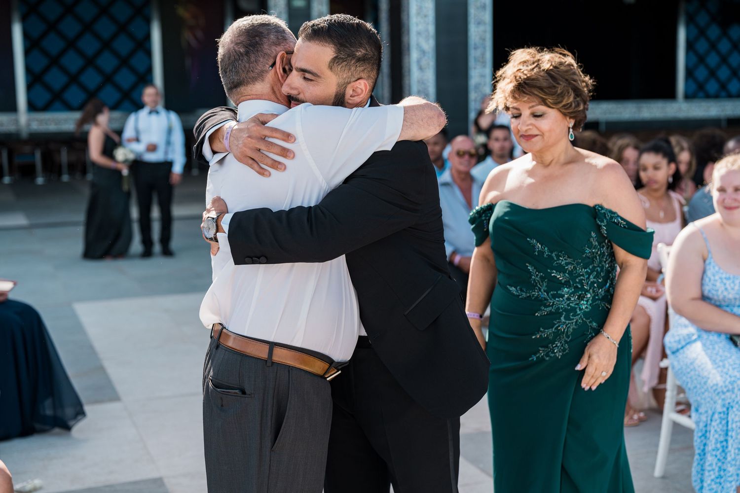 Groom embracing his father emotionally during a wedding reception while mother watches in elegant green gown.