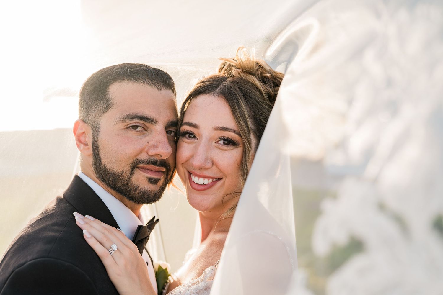 Smiling bride and groom pose closely together under a wedding veil in soft sunlight.
