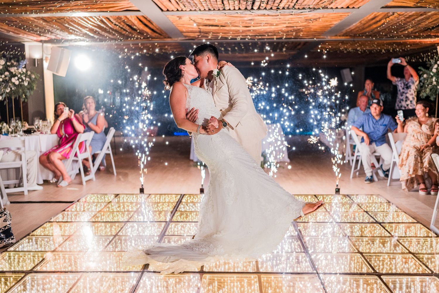 Bride and groom share first dance kiss on a mirrored dance floor with sparkling fireworks.