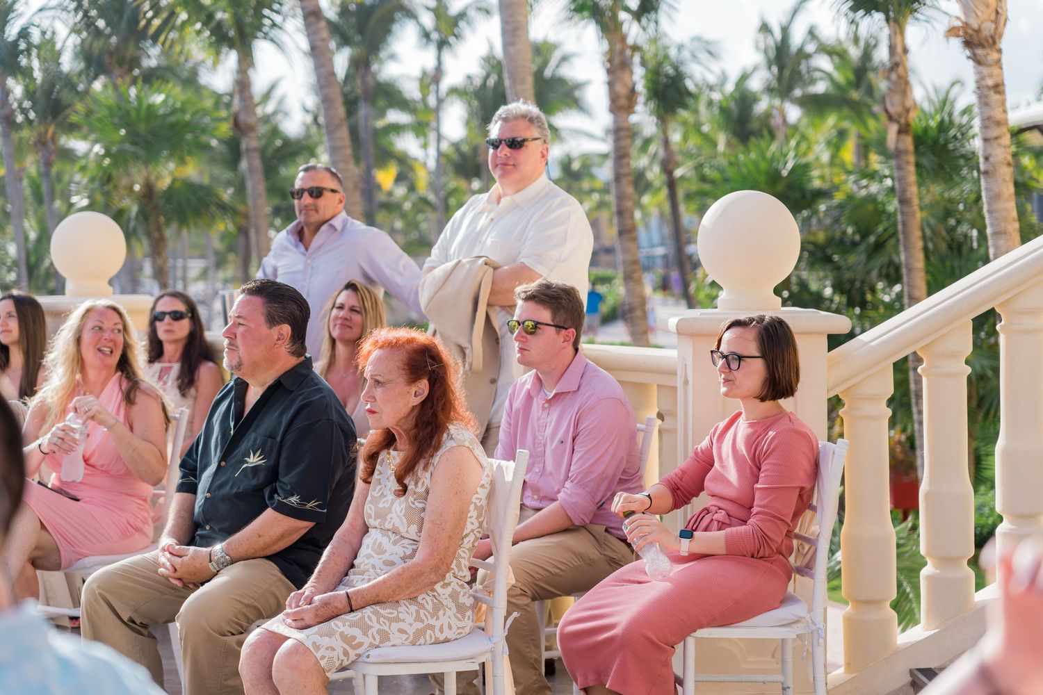 Wedding guests seated outdoors at a tropical beach ceremony with palm trees in the background.