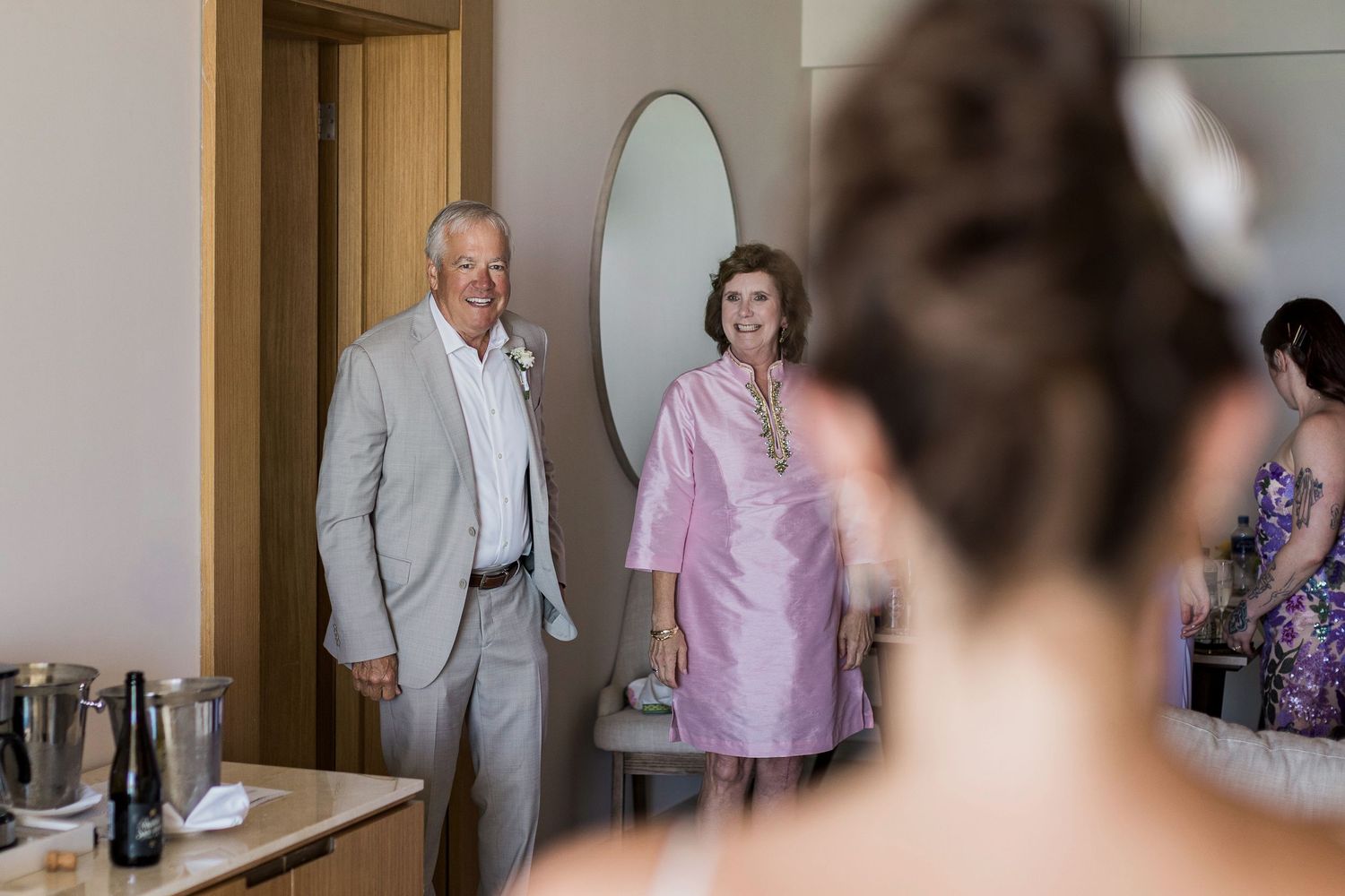 Bride seen from behind as her parents enter the room smiling during wedding preparations.