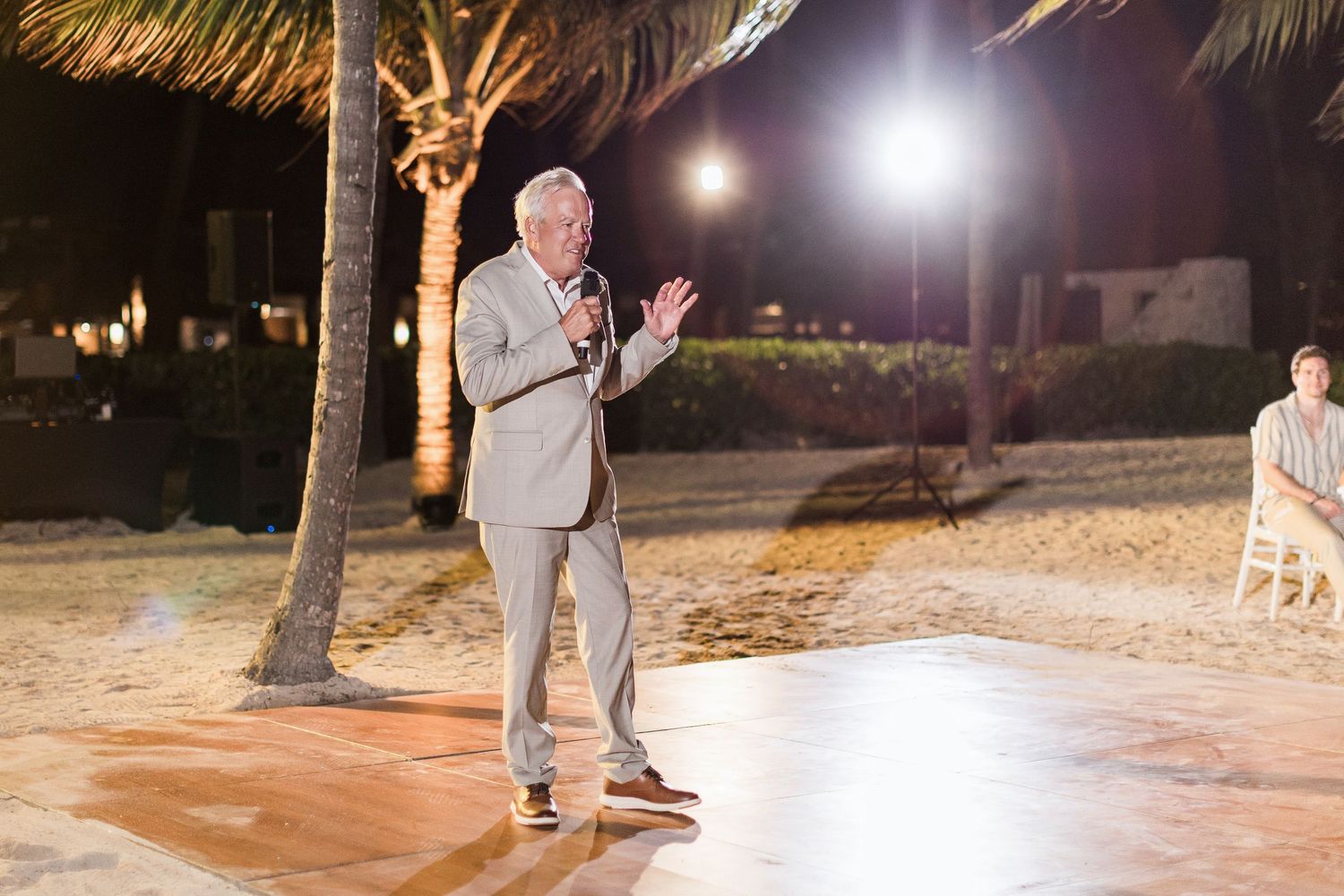 Man in beige suit speaking into microphone at outdoor beach wedding reception at night with palm trees.