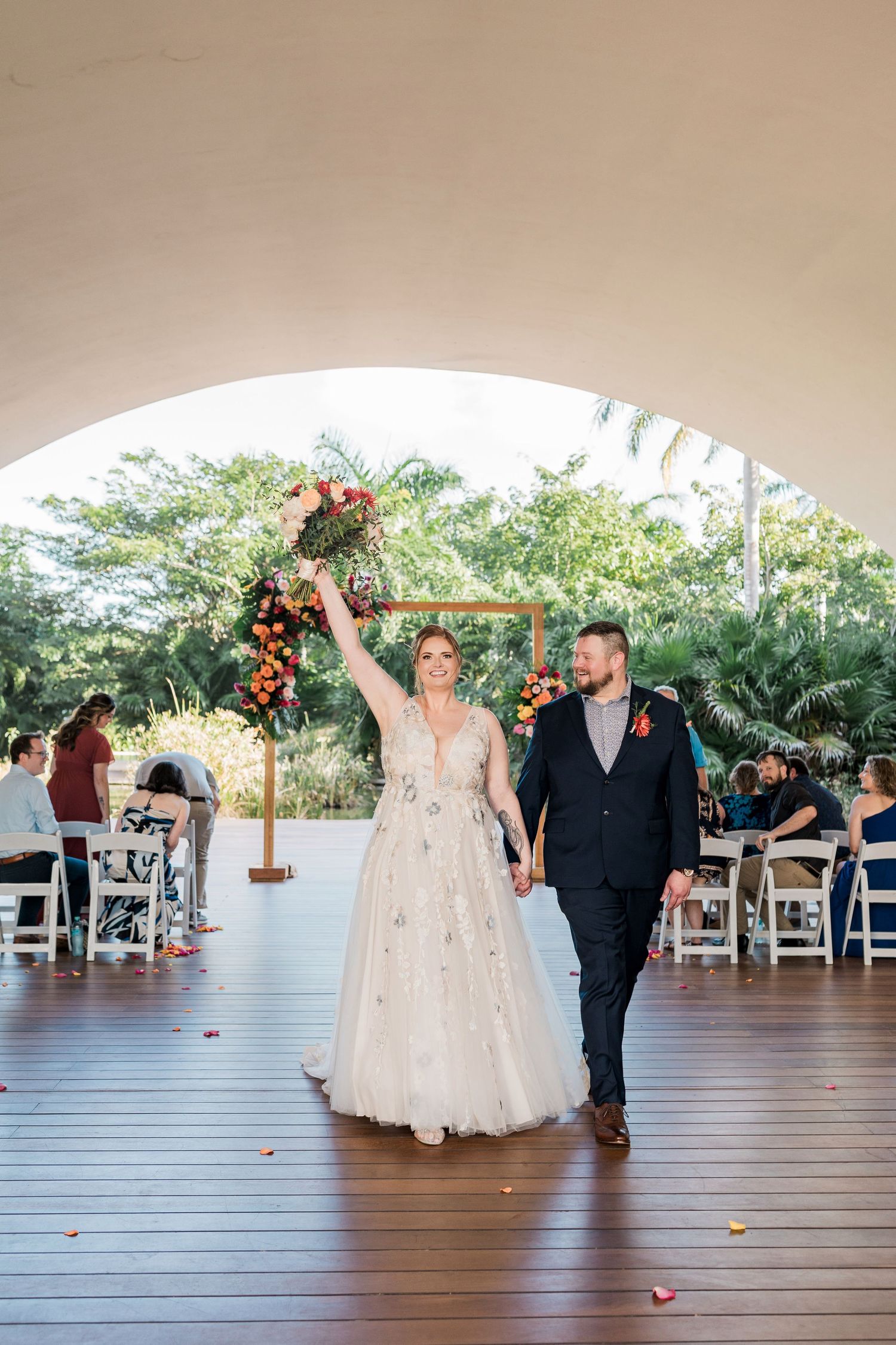 Secrets Akumal Wedding - Bride raises bouquet while walking with groom down aisle after outdoor wedding ceremony under arch.