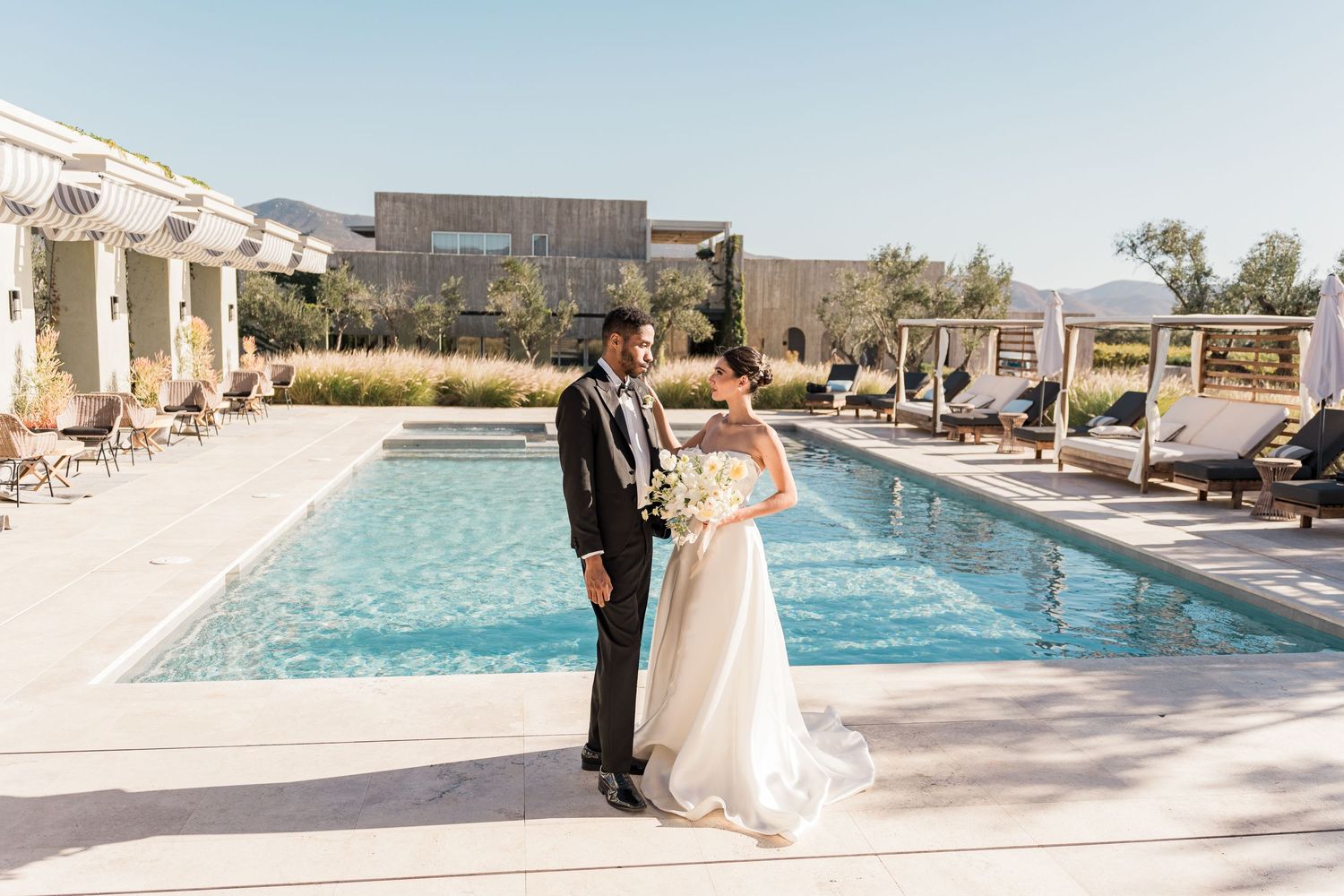 Bride and groom standing together beside a resort pool on their sunny wedding day.