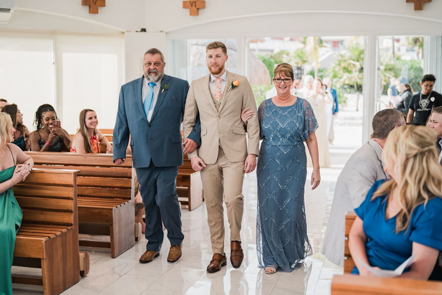 Groom walks down church aisle escorted by his parents during a bright white chapel wedding ceremony.