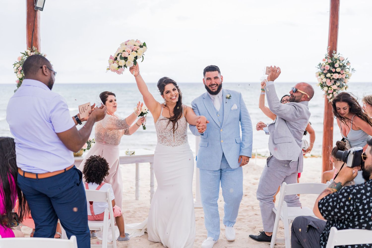 Joyful newlyweds celebrate recessional on beach as guests cheer, bride raises bouquet triumphantly.