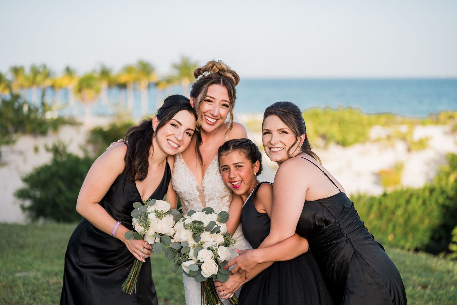 Bride and three bridesmaids in black dresses laugh and hug together with tropical beach backdrop.