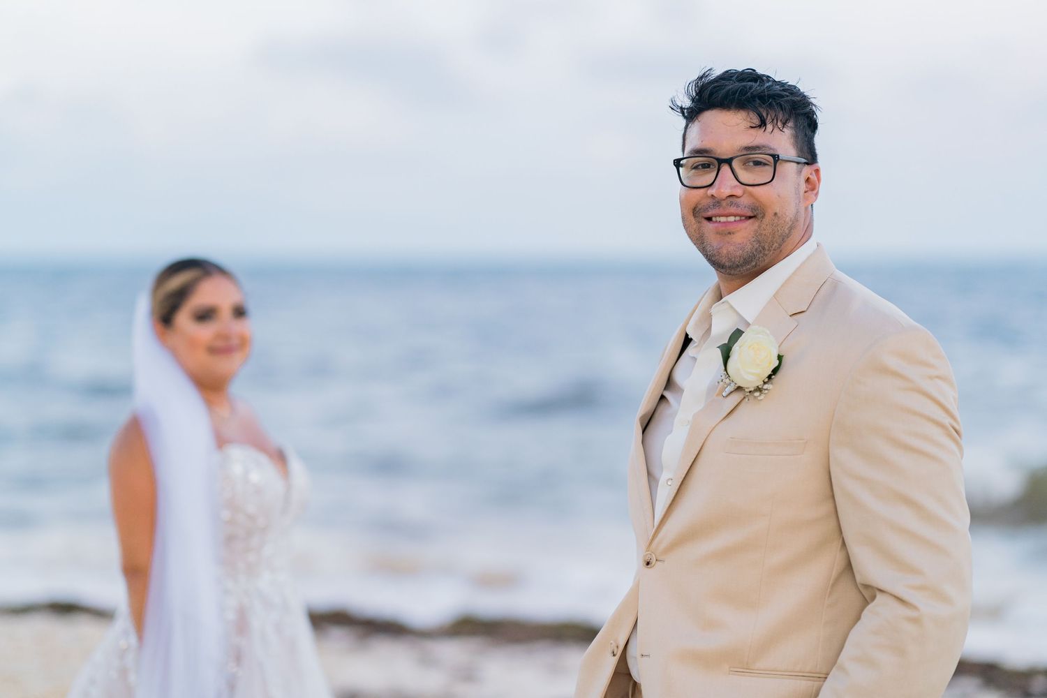 Groom in beige suit smiling at camera with bride in white wedding dress and veil blurred in background on beach.