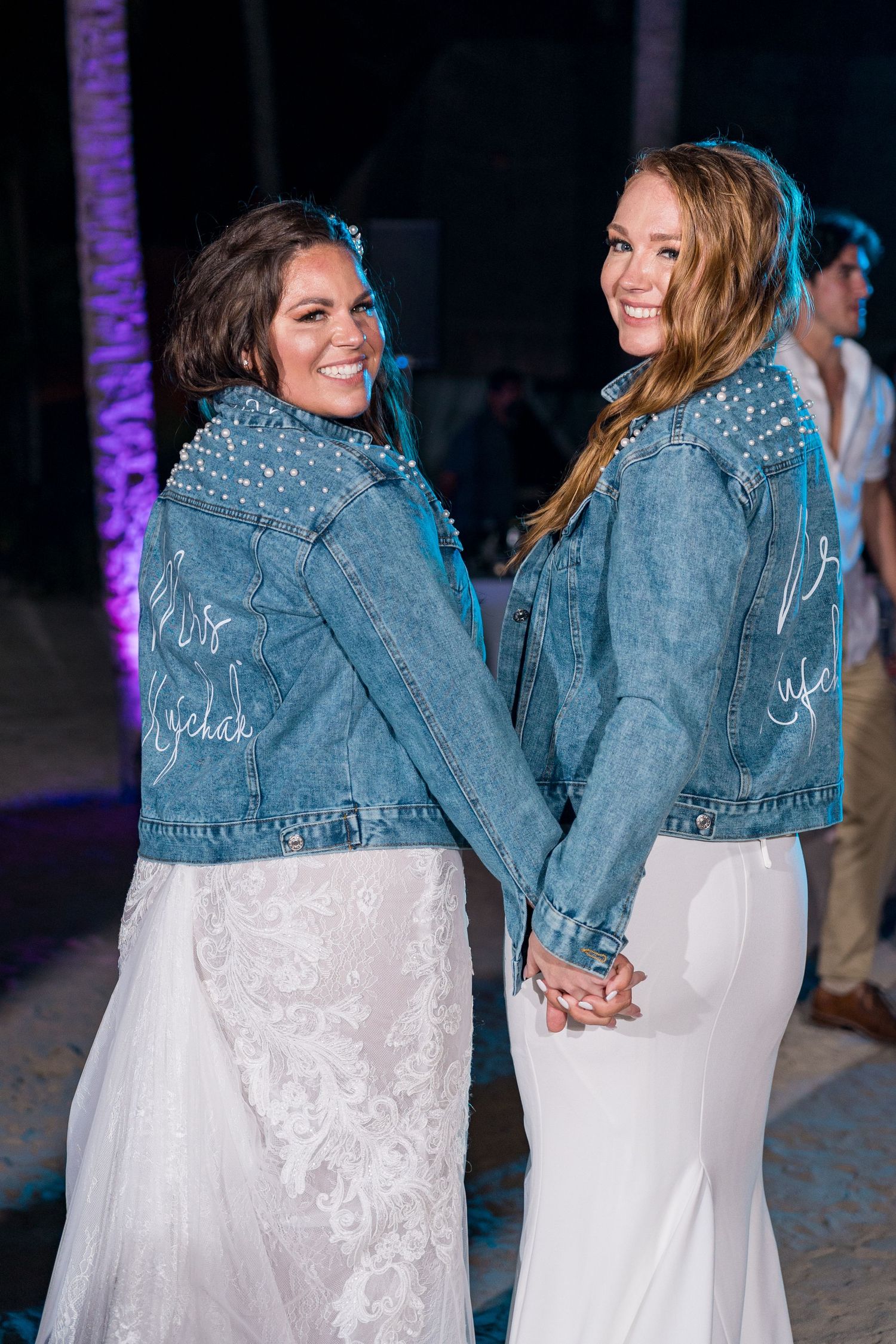 Two brides smiling in studded denim jackets and white wedding gowns at beach reception.