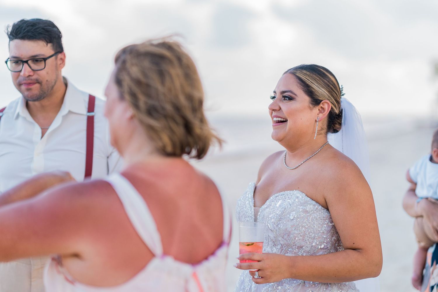 Bride in beaded gown holds a drink and smiles while chatting with guests at a beach wedding reception.