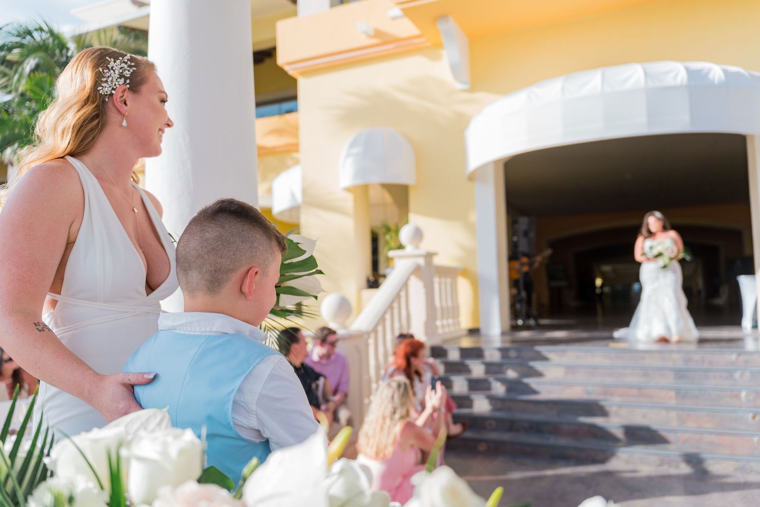 Bride and young boy watching another bride approach during an outdoor tropical wedding ceremony.