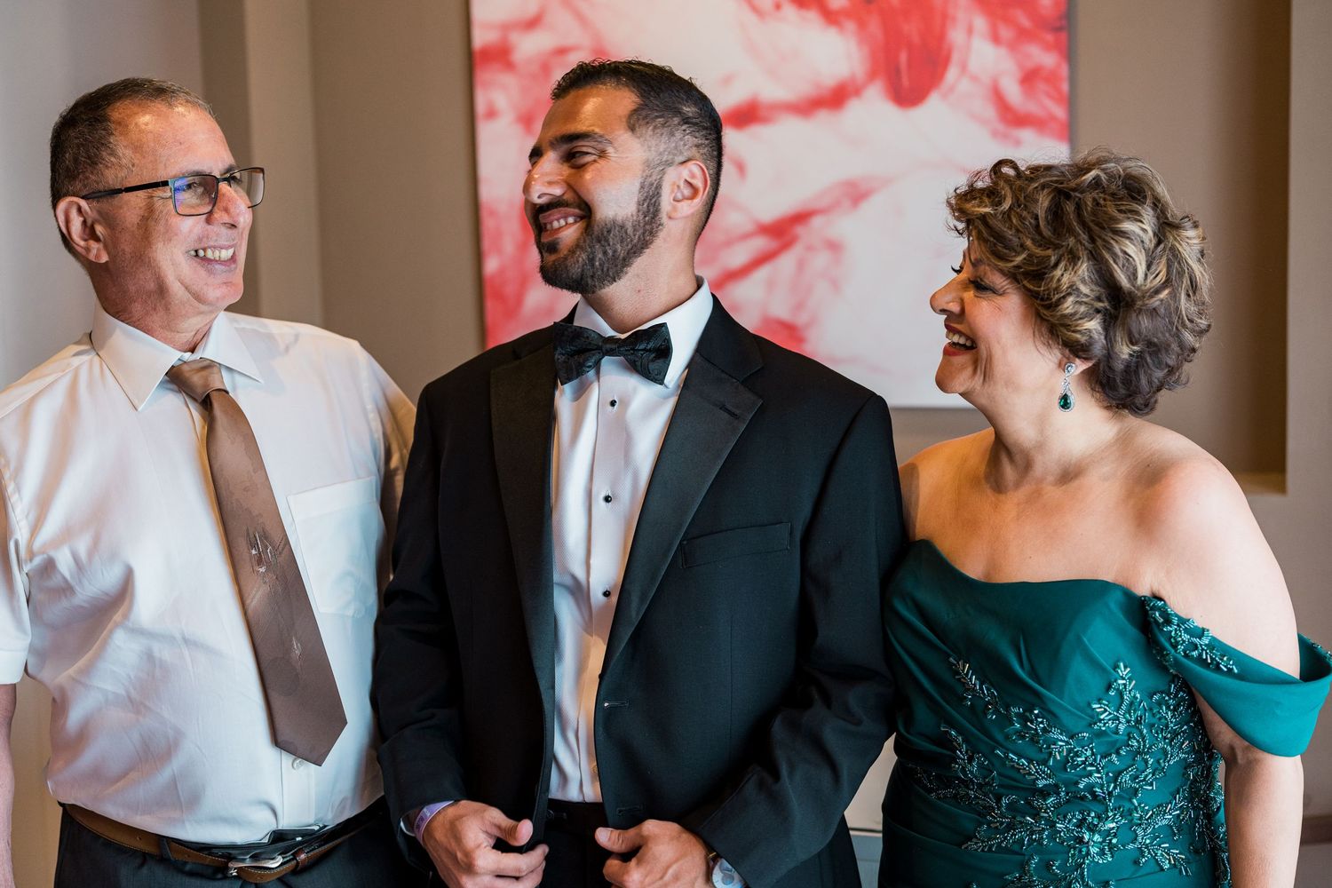 Groom in black tuxedo shares joyful moment with parents before wedding ceremony.