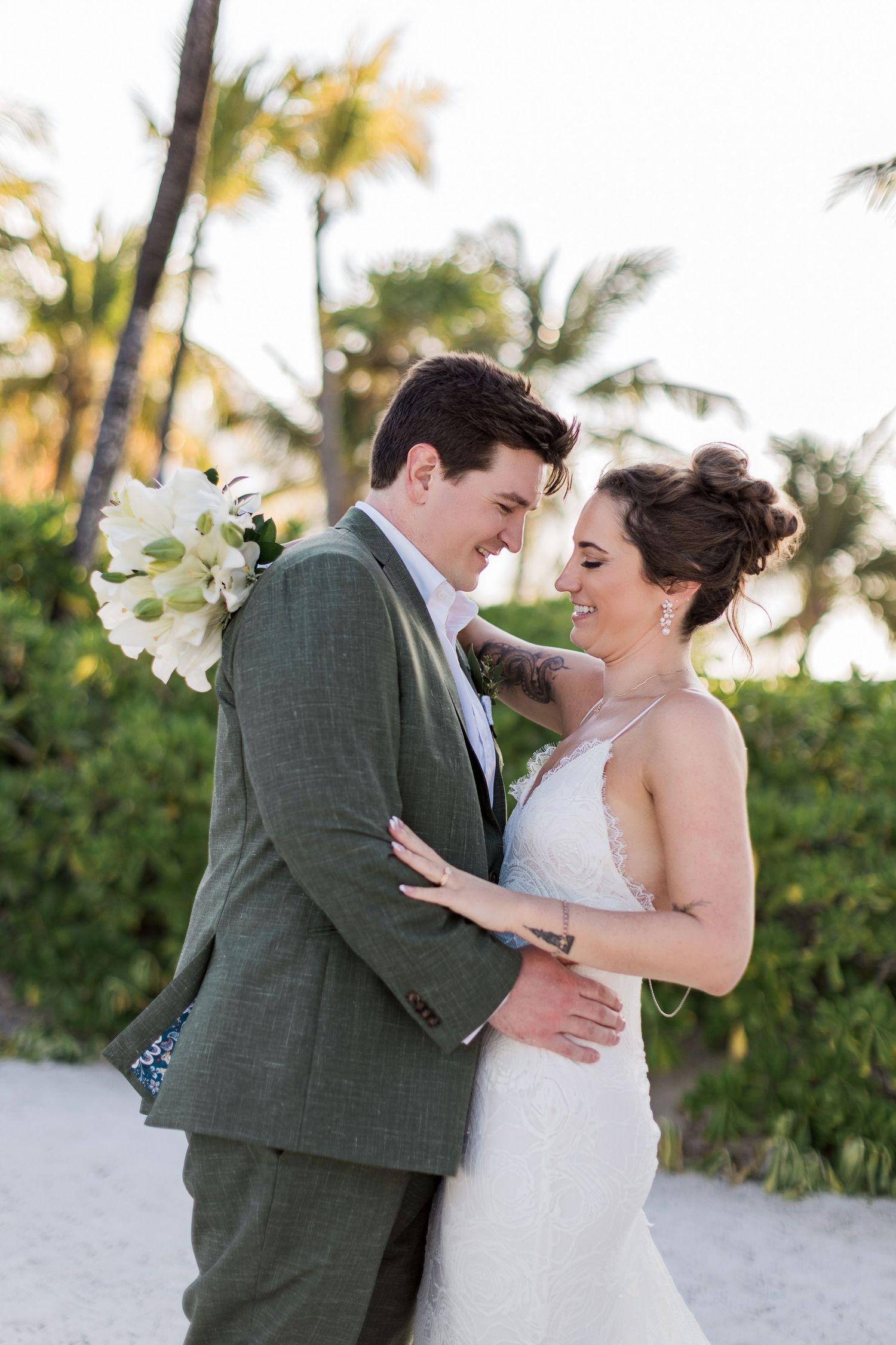 A smiling bride and groom embrace on a tropical beach, she in a white gown, he in a green suit holding white flowers.