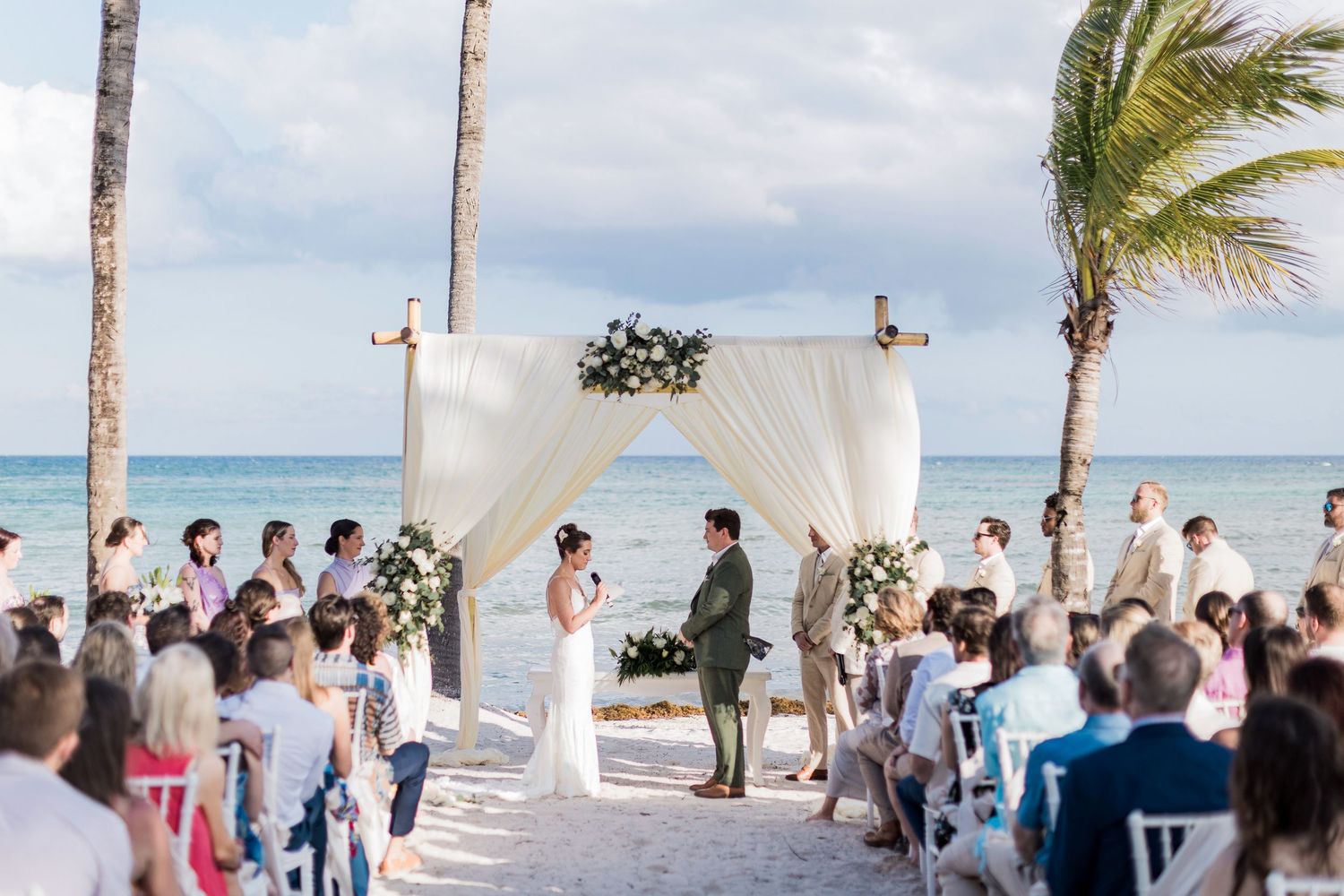 Romantic beach wedding ceremony with white draped arch, floral arrangements, and guests seated on white chairs by the ocean.