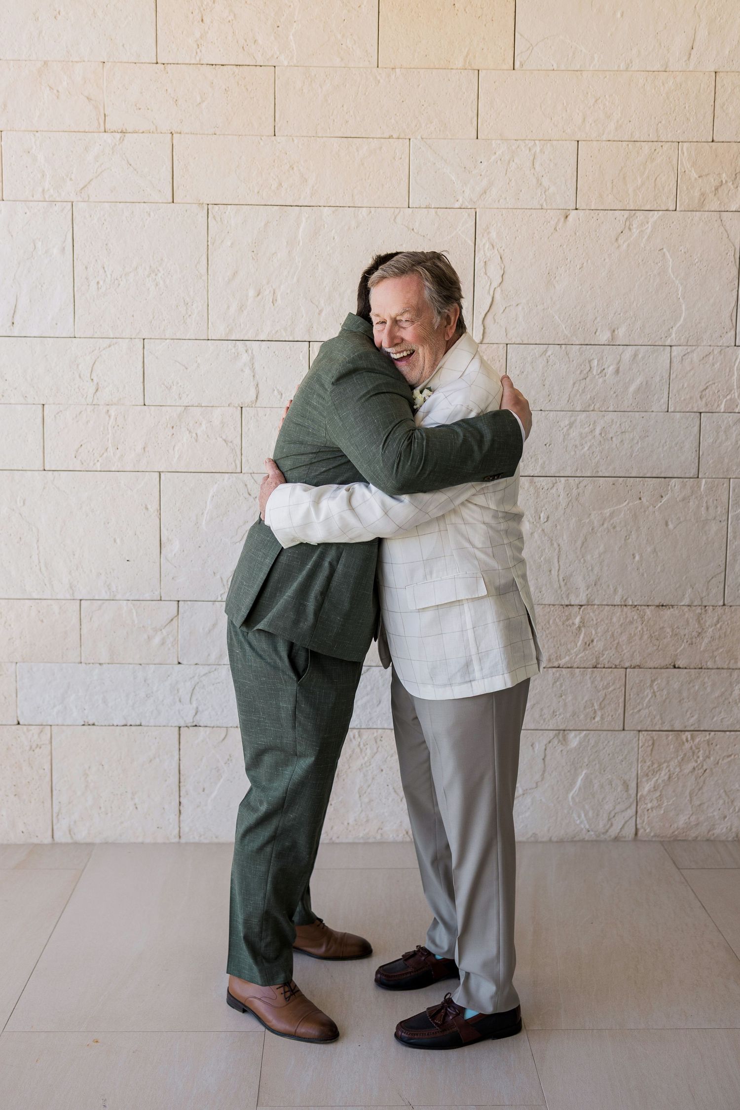 Two men in formal suits share a warm embrace while laughing against a textured white stone wall.