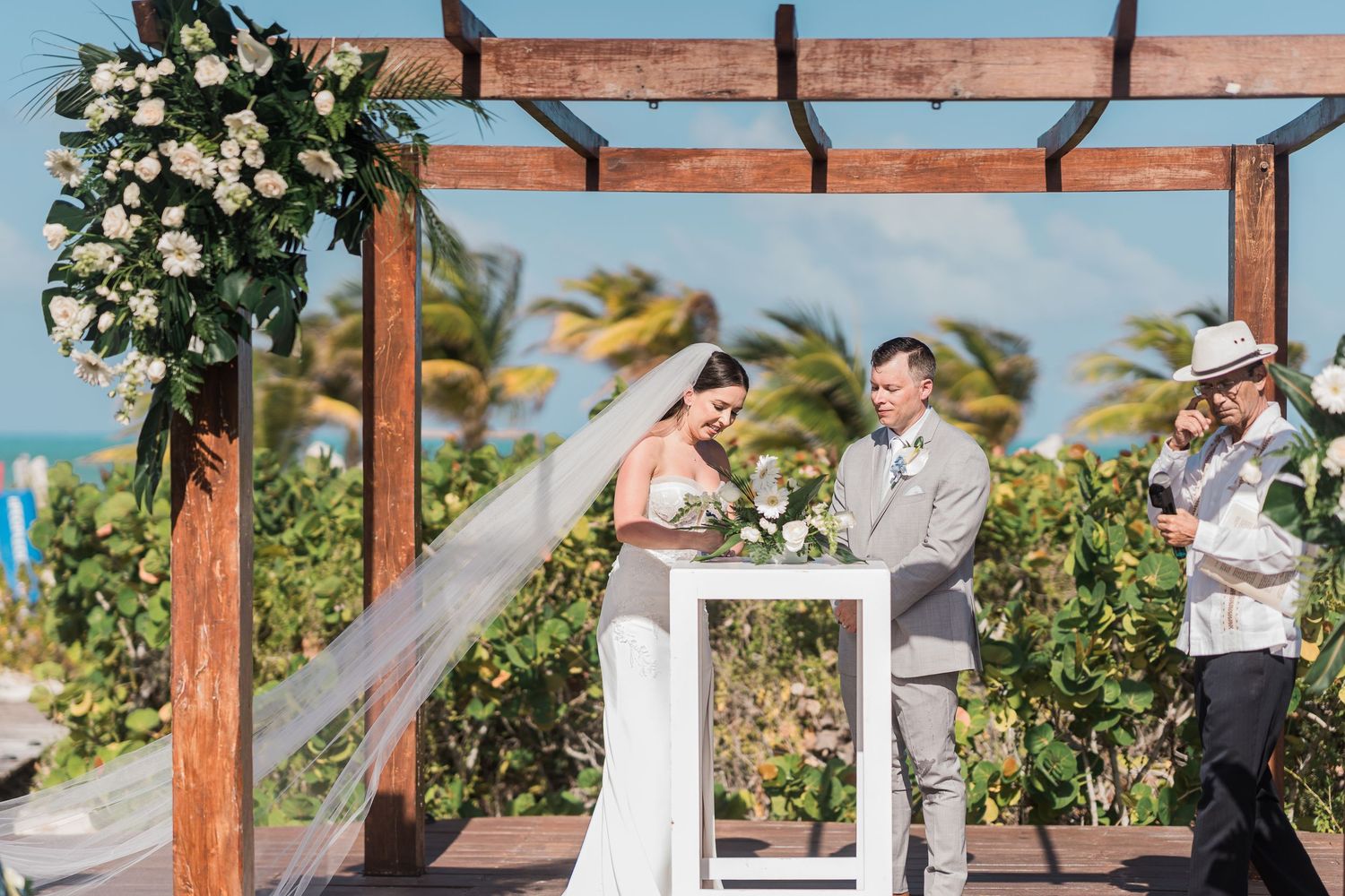 Bride and groom exchange vows at outdoor beachfront wedding ceremony under wooden pergola with tropical floral arch.