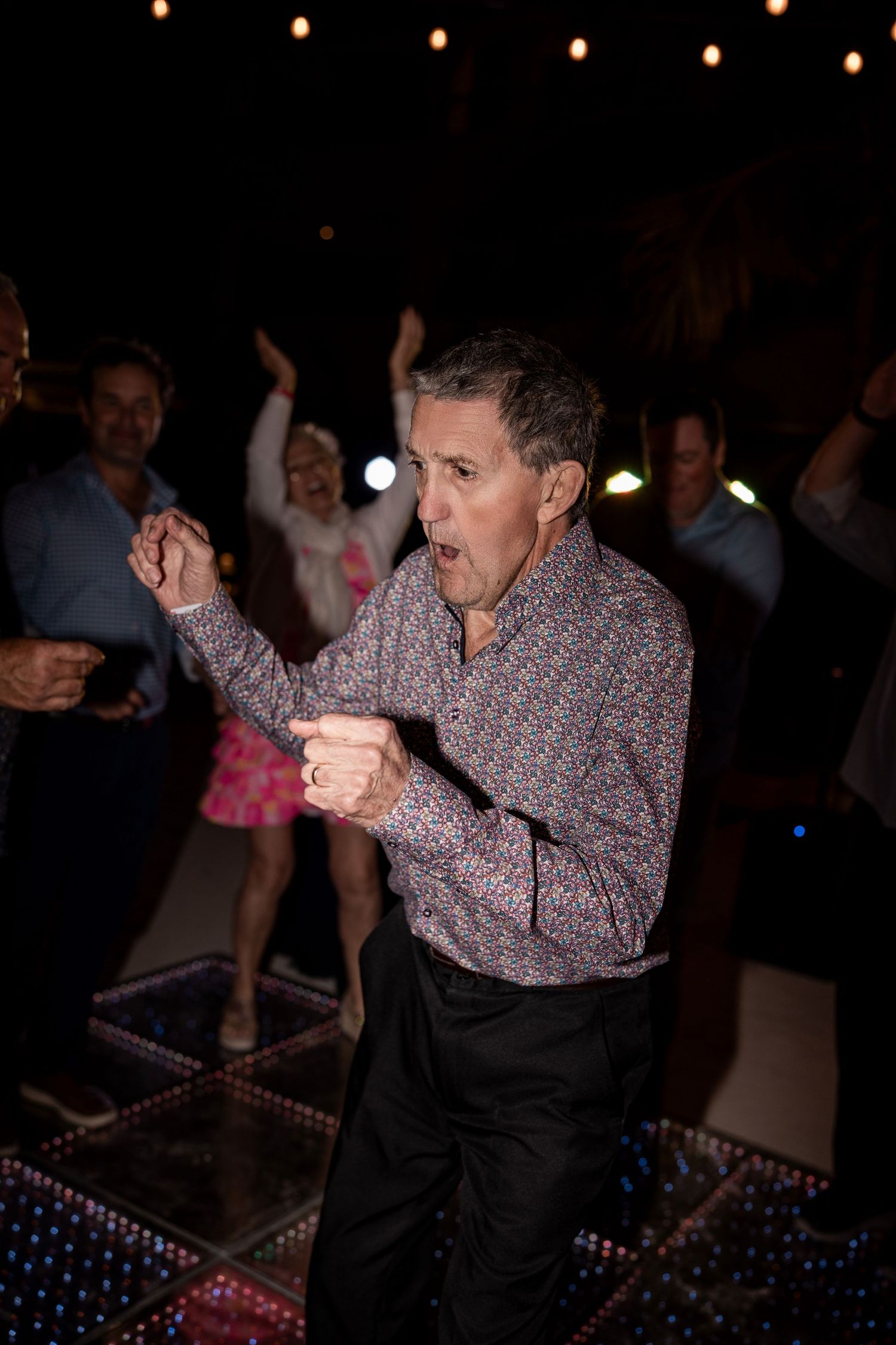 Energetic man dancing at a party on a lit dance floor, wearing a floral shirt, with guests celebrating in the background.