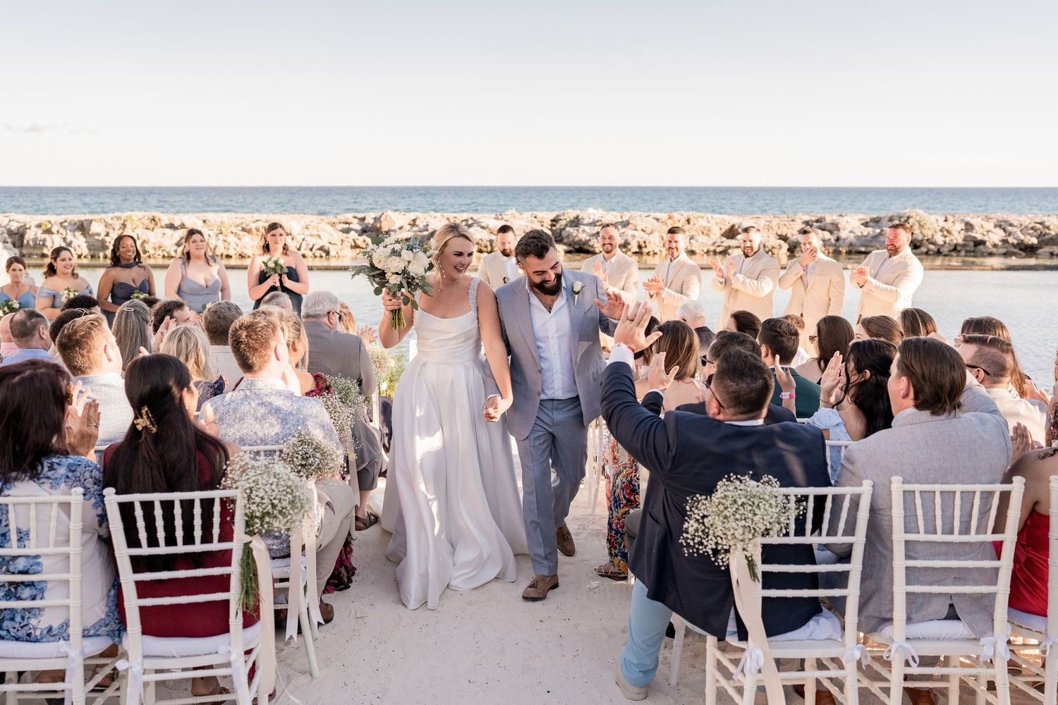 Bride and groom walk down aisle at outdoor beach wedding ceremony with guests cheering.